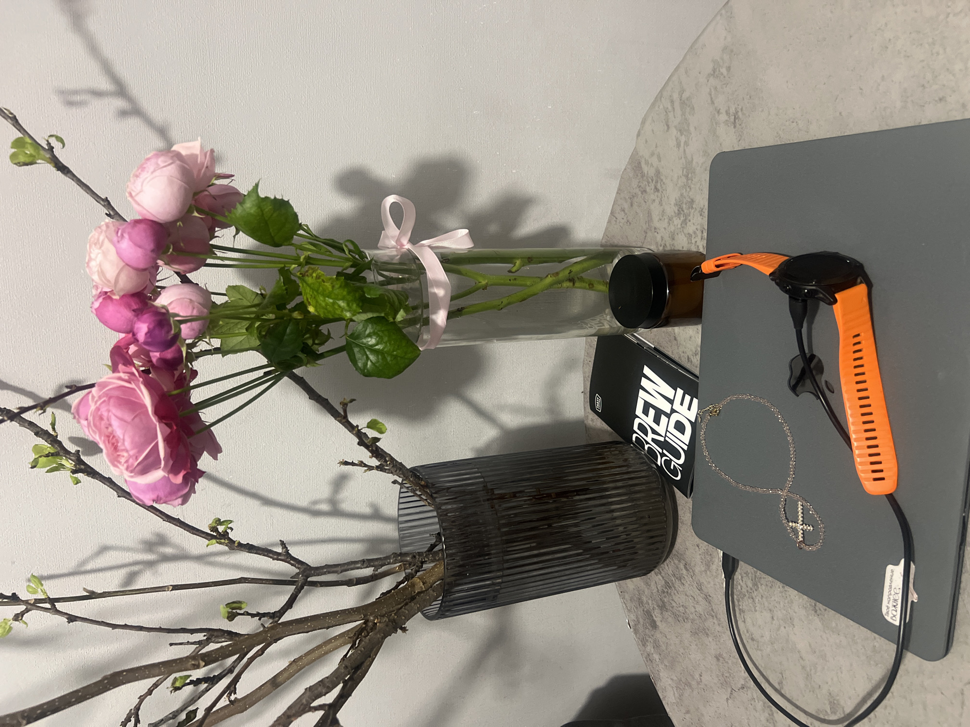 The image depicts a tabletop scene, likely indoors, featuring a laptop, vases with flowers, and various accessories.
A grey laptop, possibly a MacBook, is positioned on the right side of the frame, with an orange smartwatch placed on top. A charging cable is connected to the watch and runs along the side of the laptop. A silver necklace with a cross pendant is also draped on the laptop.
To the left of the laptop, there are two vases of flowers. One vase is a clear glass tube tied with a pink ribbon, containing pink roses and greenery. The other vase is a dark, ribbed glass container holding what appears to be bare branches with small buds.
Between the vases and the laptop, a black booklet labeled "BREW GUIDE" is visible.
The background is a light grey wall, and the tabletop appears to have a speckled grey and white pattern.
There is no information provided in the image that relates to the city of Kazan, Russia.