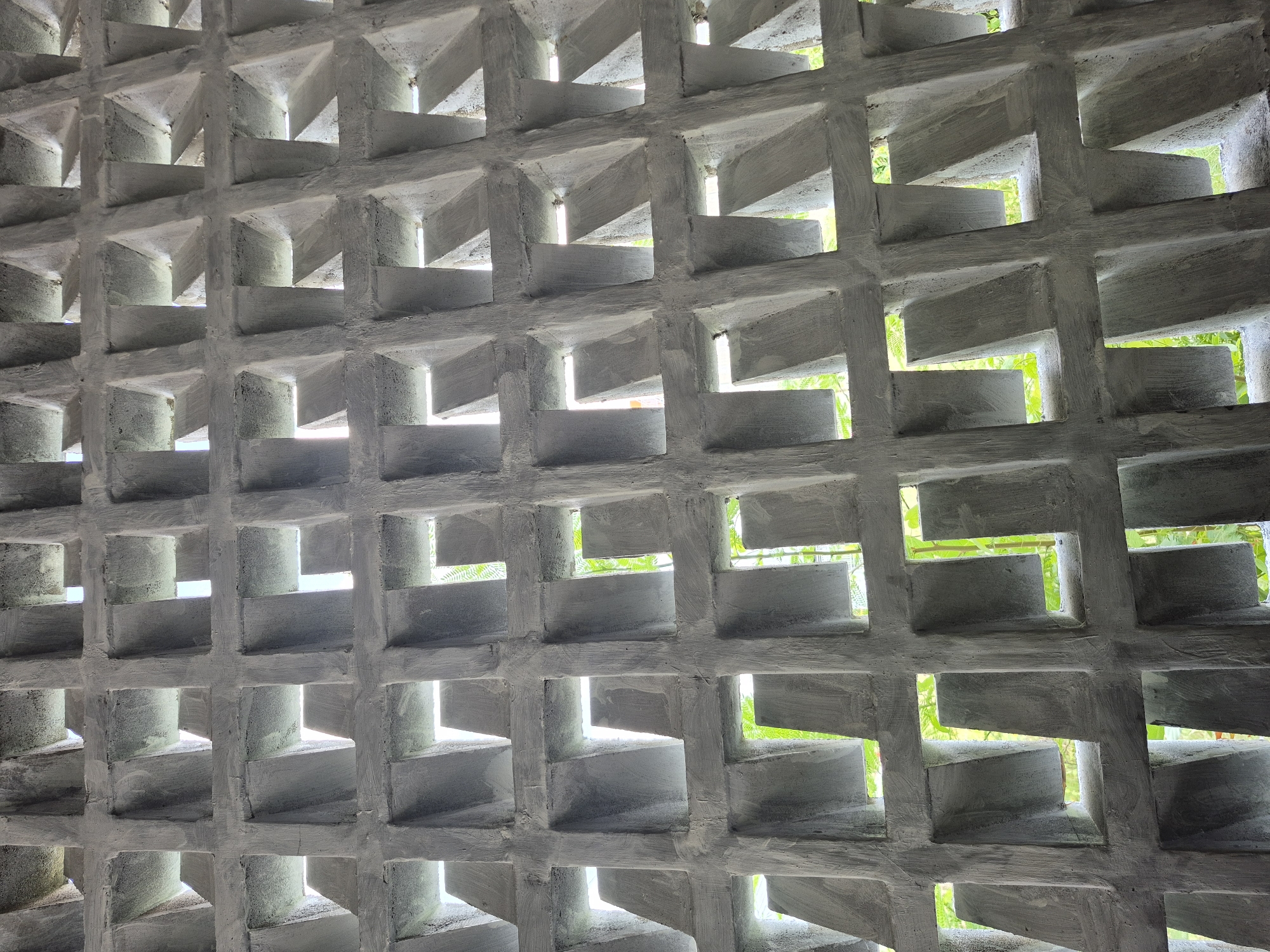 An image captures a detailed view of a grey concrete ventilation block wall, likely serving as a facade or partition. The wall is composed of numerous square modules arranged in a grid pattern. Each module features a complex, three-dimensional internal structure formed by L-shaped concrete elements, creating angled openings that allow light and air to pass through while providing some visual obstruction. This design creates a dynamic interplay of light and shadow across the rough, textured surface of the concrete, highlighting the architectural relief.
Through the various openings, bright green foliage is visible, suggesting an outdoor setting or a lush garden immediately beyond the wall. The strong sunlight filtering through the blocks casts distinct shadows and creates bright highlights on the edges of the concrete, indicating it is daytime. The overall impression is one of modern tropical architecture, designed for natural ventilation and sun shading, a common feature in climates like Lhokseumawe, Indonesia. There are no people, explicit activities, or visible text in the image.