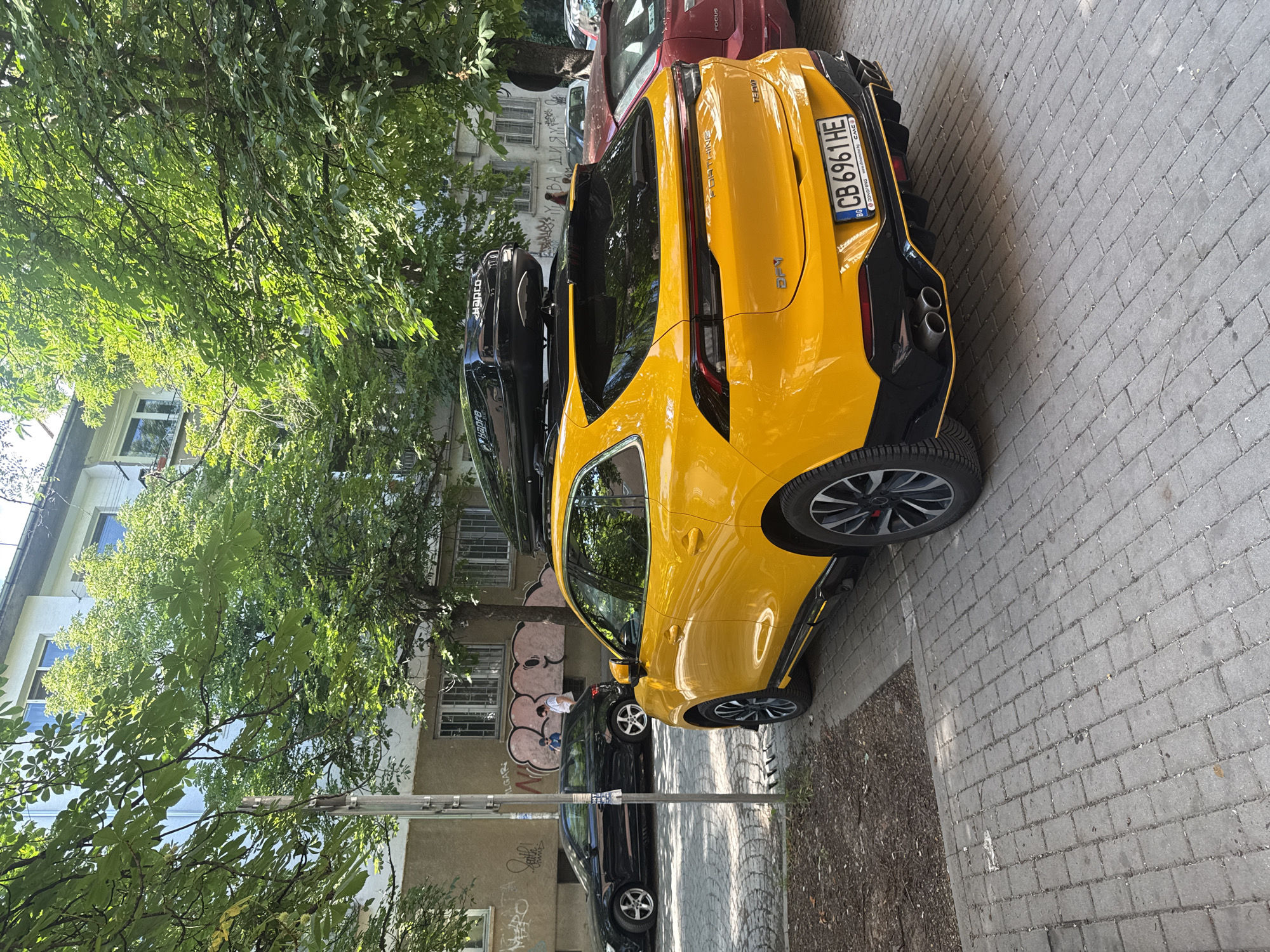 The image shows a bright yellow car parked on a cobblestone street in Sofia, Bulgaria. The car has a black roof box attached to its roof. The car is a modern SUV with a sleek design, featuring sharp lines and aggressive styling. The license plate of the car reads "CB 6961 HE". In the background, there is a building with white and beige facades, and trees with lush green foliage. Other cars are parked on the street, including a black Audi and a red car. There are also a couple of people visible in the background, near the building. One person is wearing a blue shirt and another is wearing a white shirt. Graffiti is visible on the wall of the building. The scene appears to be a typical urban environment in Sofia.