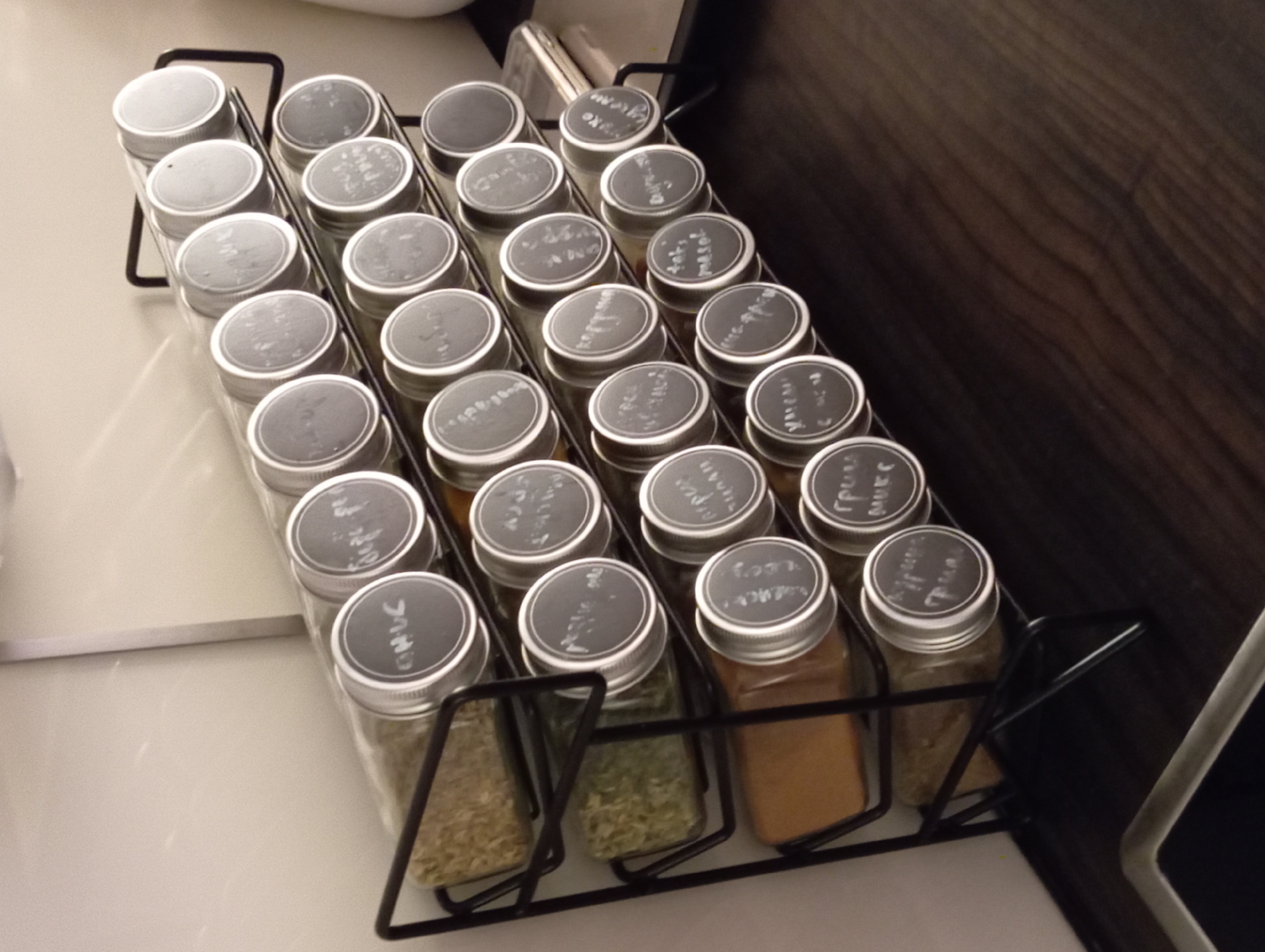 This image shows a black wire spice rack with four shelves filled with glass jars of spices. The jars have silver lids with round black labels written on in white chalk. The spice rack is positioned on a dark brown wooden countertop, likely in a kitchen. The background is a light-colored wall. The image is taken from a slightly elevated angle, looking down and across the spice rack. It is unclear if this image is located in Baden, Austria.