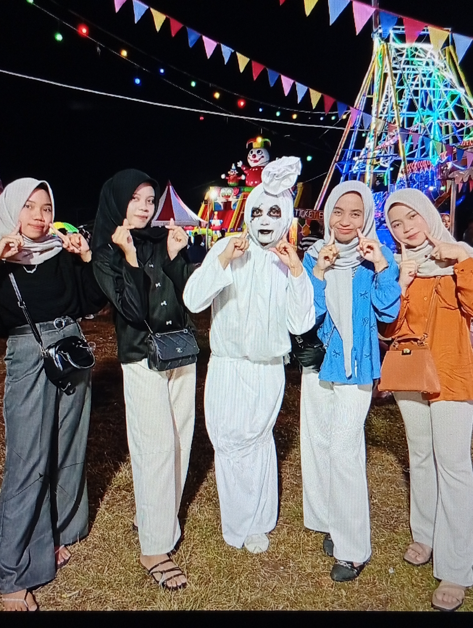 The image displays five individuals, predominantly young women, posing outdoors at night in a fairground setting located in Lhokseumawe, Indonesia. The ground consists of dry, matted grass. The background features various carnival attractions: a large Ferris wheel-like structure illuminated with multicolored lights (blue, green, yellow, pink) on the right, a red and white striped tent or booth, and a large red and white clown figure. Colorful triangular bunting and string lights are strung across the dark night sky. A partially visible sign reads "TICKET S".
From left to right:
1. A young woman wearing a light grey hijab, a black long-sleeved shirt, dark grey wide-leg trousers, and light-colored flip-flops. She carries a black crossbody bag with a silver chain strap and forms a finger heart gesture near her left cheek.
2. A young woman wearing a black hijab, a black long-sleeved shirt, light beige wide-leg trousers, and dark sandals. She carries a black quilted crossbody bag with a silver chain strap and forms a finger heart gesture with both hands.
3. An individual in an all-white costume, featuring a white headpiece, long-sleeved top, and wide-leg trousers. The face is painted white with dark outlines around the eyes, lips, and extending from the mouth. They form finger heart gestures with both hands near their face and wear light-colored closed-toe shoes.
4. A young woman wearing a light grey hijab, a blue long-sleeved top with white embroidered patterns, and white wide-leg trousers. She forms finger heart gestures with both hands near her cheeks and wears black embellished flat shoes.
5. A young woman wearing a light beige hijab, an orange long-sleeved top, and light beige wide-leg trousers. She carries a light brown structured handbag and forms a finger heart gesture with both hands near her chin.
All visible individuals are looking towards the camera.