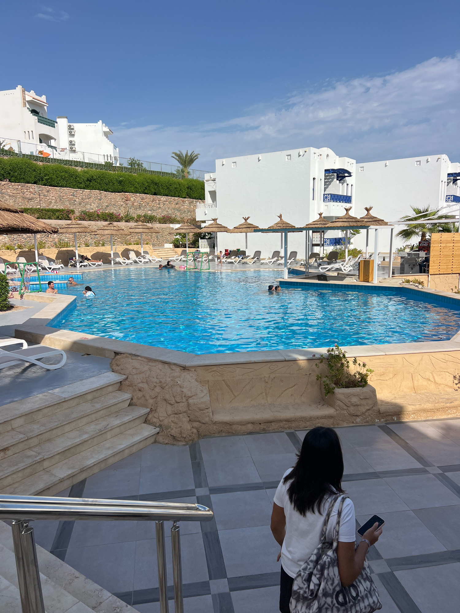 The image captures a poolside scene at a resort in Sharm El Sheikh, Egypt. A large, bright blue swimming pool dominates the center of the frame, with several people swimming and relaxing in the water. The pool is surrounded by a textured, light brown border and a paved patio area. Lounge chairs and straw umbrellas are set up around the pool for sunbathers. In the background, white resort buildings with blue accents are visible, built on a terraced hillside covered with lush green vegetation. A clear blue sky with scattered clouds completes the serene atmosphere of a sunny day.