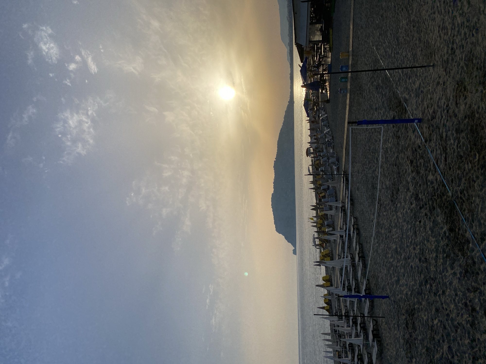 This image depicts a beach scene in Alanya, Turkey, at sunset. Rows of sunbeds and umbrellas line the sandy shore, stretching towards the sea. The sun is positioned in the upper center, casting a golden glow across the sky and water. A mountain range is visible in the distance. In the foreground, there is a volleyball net on the sand. The sky is partly cloudy. There are no people visible in the image.
