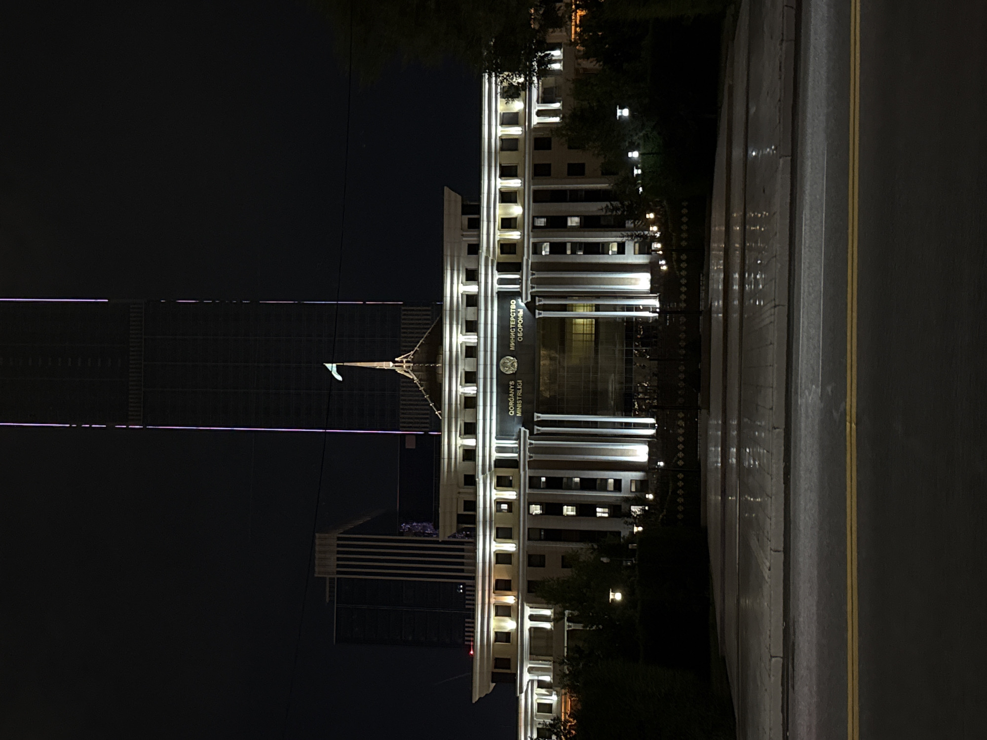 The image captures the Ministry of Defense building in Astana, Kazakhstan, at night. The building is illuminated by warm white lights, highlighting its neoclassical architecture with columns and a grand entrance. Behind the Ministry, a tall skyscraper stands out with its brightly lit facade, contrasting with the darker sky. In front of the building, a wet paved area reflects the lights, suggesting recent rain. The foreground shows a road with yellow double lines, indicating a street in the city. The scene is devoid of people, presenting a quiet and imposing nocturnal view of a significant government structure.