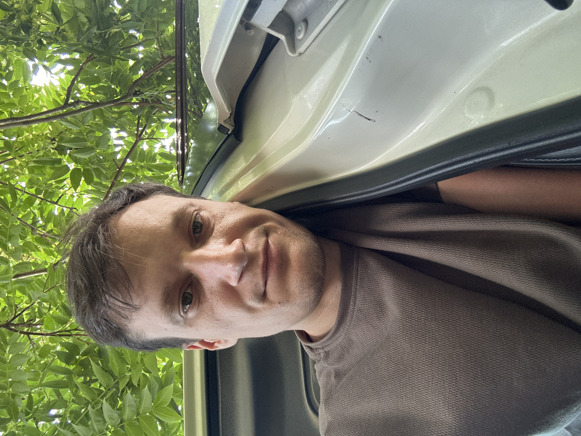 A man, appearing to be in his late 20s or early 30s, takes a selfie outdoors. He is positioned leaning against the open door of a light-colored vehicle, with his head resting on the car's frame. The backdrop consists of lush green foliage from overhanging trees, suggesting a park or shaded area. The man is wearing a brown, textured t-shirt. The lighting indicates it's daytime. The image focuses on the man's face and upper body, with a portion of the car's interior and exterior visible. The specific location is a street in Makiv, Ukraine.