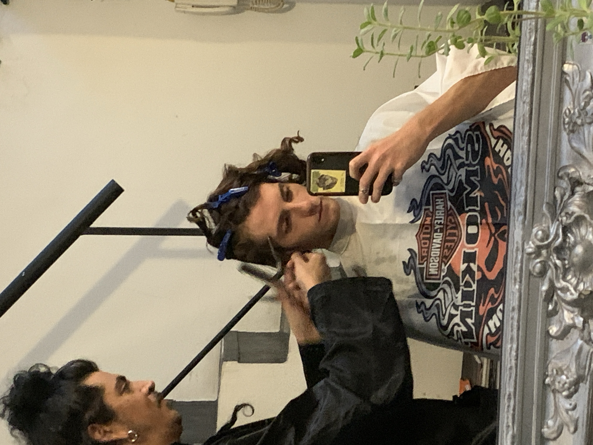 A mirrored reflection shows two individuals. One person, facing to the left, has dark, curly hair, a mustache, and wears an earring. They are draped in a black garment and appear to be holding scissors, as if cutting the hair of the other individual. The other person, whose reflection is more central, is taking a selfie using a smartphone. Their hair is partly clipped up with blue hair clips. They wear a white shirt with a prominent logo featuring the words "OKIMA" and "HARLEY-DAVIDSON." The location appears to be indoors, possibly a home or a small salon. Buenos Aires, Argentina, is not discernible from the image alone. The background includes a white wall, part of a staircase, a plant, and an ornate silver frame of the mirror.