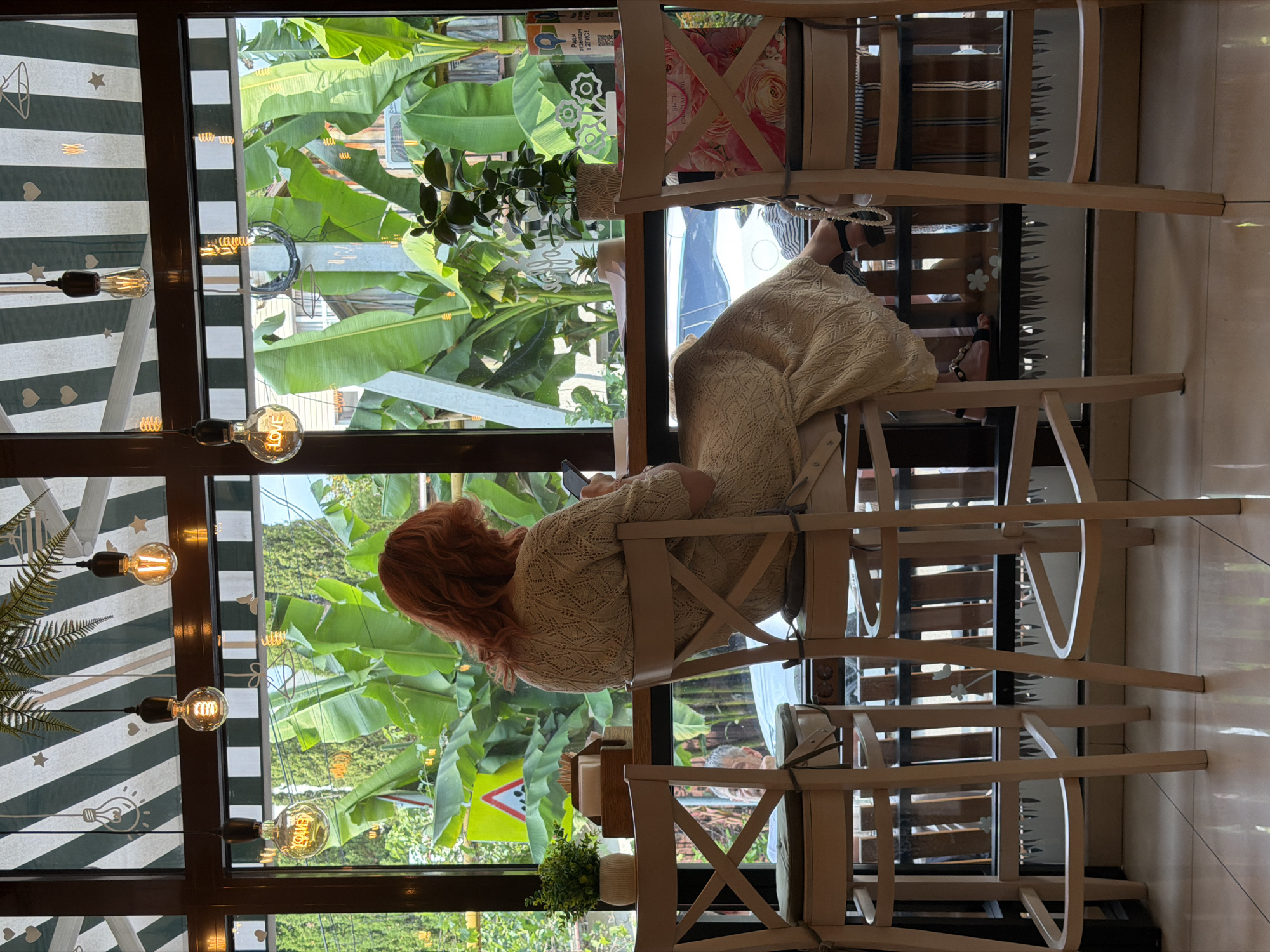 A woman with red hair sits at a high chair in a cafe, facing a window that overlooks a lush green outdoor area with large banana leaves. She is dressed in a cream-colored, knitted dress and is looking at her phone. The cafe interior features three hanging Edison lightbulbs, two of which are illuminated and one of which has the word "LOVE" etched into it. The chairs are made of light wood with white cross-back detailing. On the table next to her is a small potted plant and a wooden dispenser of napkins. The scene suggests a relaxed atmosphere in a cafe in Sochi, Russia, with the bright greenery outside creating a tropical ambiance.