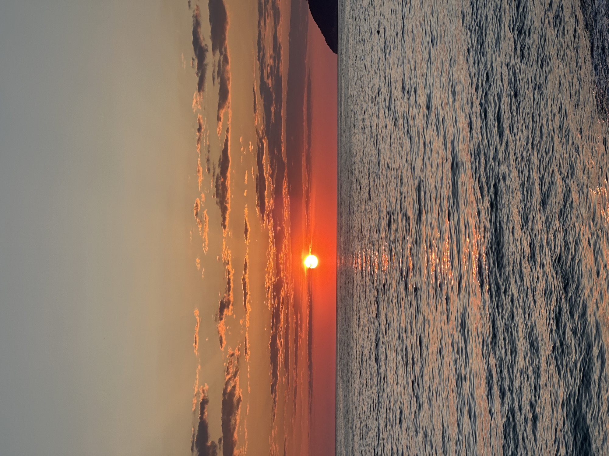 The image shows a sunset over the Adriatic Sea near Šušanj, Montenegro. The sky is partially cloudy with the sun setting in the center of the frame. The water is slightly choppy, reflecting the sunlight. The overall tone is warm and serene.
