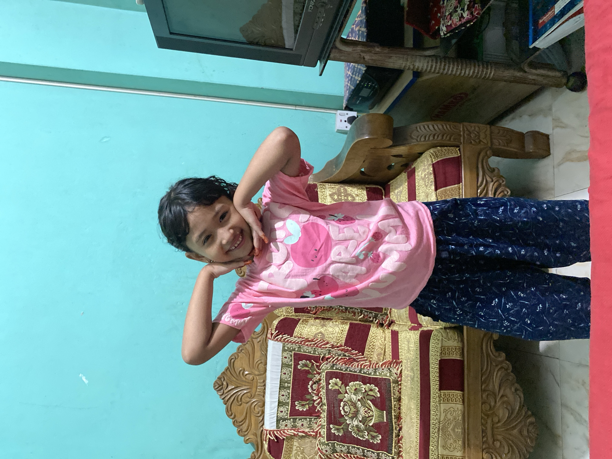A young girl is posing while seated on an ornate sofa in what appears to be an indoor setting in Dhaka, Bangladesh. She is wearing a pink T-shirt with a graphic print and dark blue patterned pajama pants. The girl has dark hair and is smiling, holding her hands up to her cheeks in a playful gesture. The sofa has a wooden frame with detailed carvings and is upholstered with a patterned fabric in shades of gold, burgundy, and white. A turquoise-colored wall is visible in the background. Additionally, an old television set and some items are visible in the background.