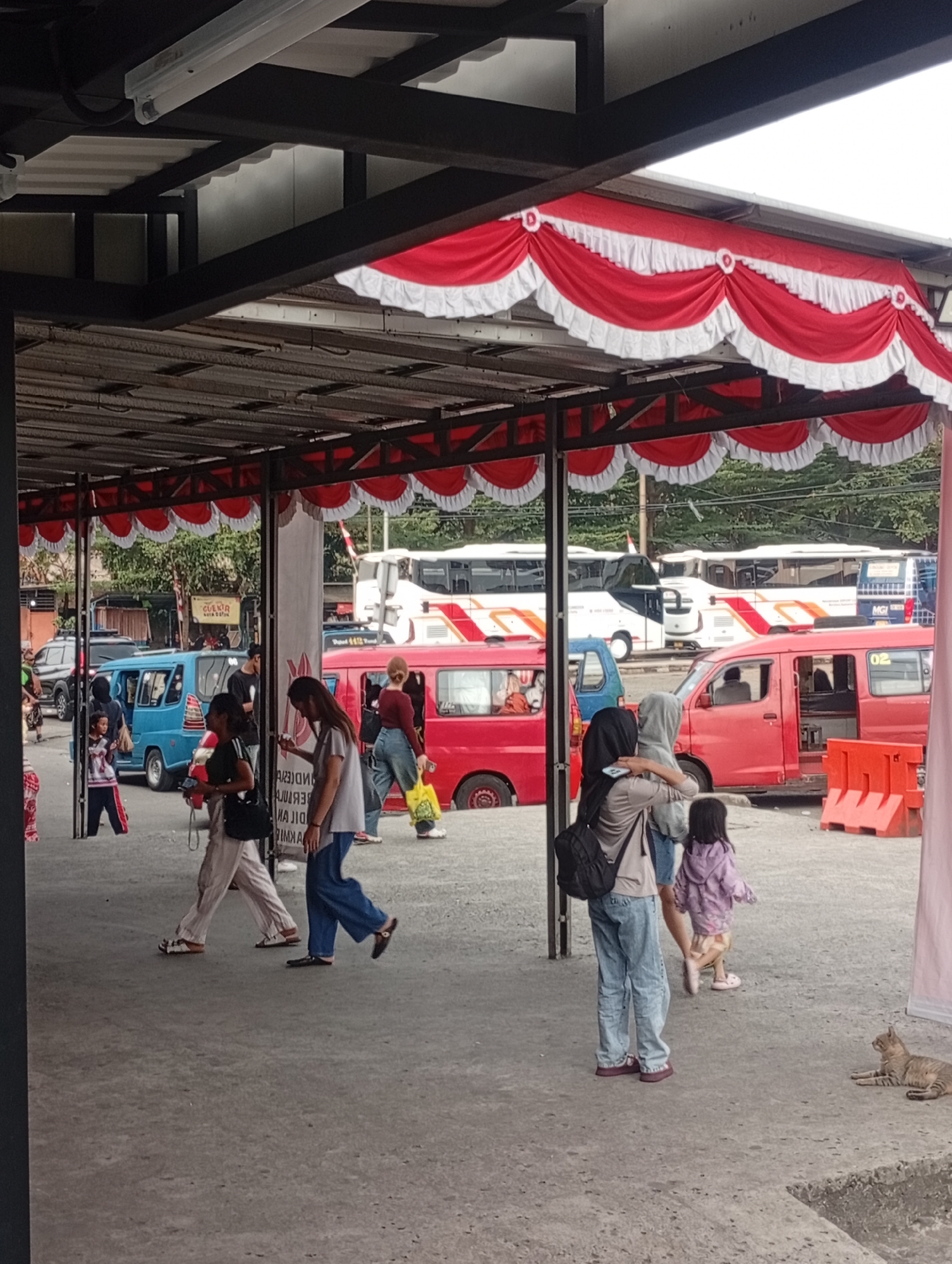 The image depicts a bus terminal or waiting area in Depok, Indonesia, likely at the Tirta Jaya Depot. The foreground shows a concrete ground with a slight elevation change on the right side. Several people are walking and standing. A woman in the center is wearing a grey top and blue pants, walking away from the camera. To her left, a woman in a white top and light-colored pants is walking, holding a phone. Further left, a child in a striped shirt and dark pants is standing. A red minivan is parked on the left, with its door open. In the background, multiple buses are visible, parked in a lot under a covered structure adorned with red and white bunting. The environment appears to be an outdoor, covered transportation hub. A tabby cat is lying down on the concrete in the lower right corner of the image.