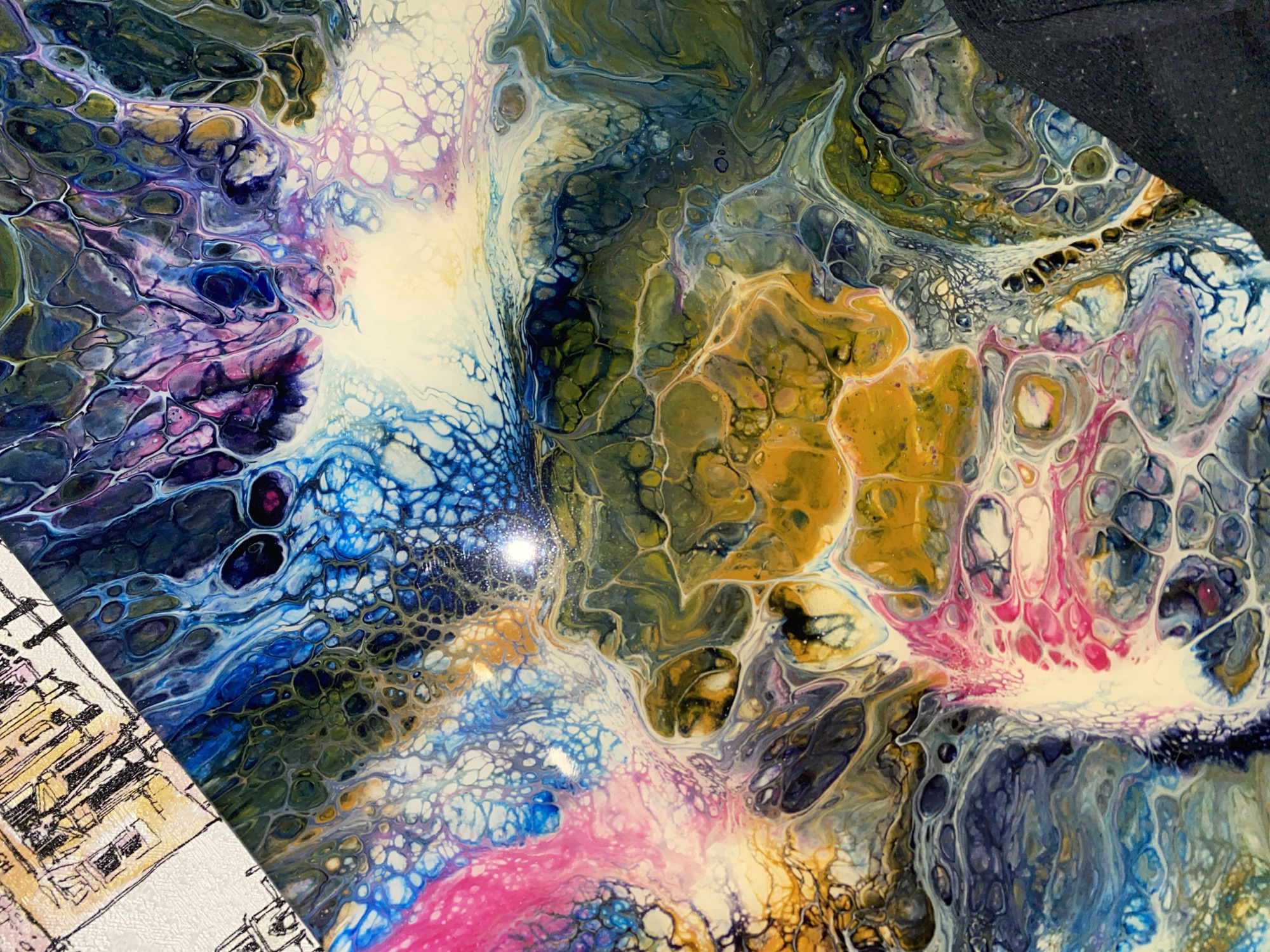 The image shows a close-up view of a pour painting. The painting features a chaotic and abstract composition of colors and textures, with vibrant hues of blue, purple, pink, yellow, and green swirling together. The technique used creates a cellular or marbled effect, with interconnected cells and organic shapes. In the upper left corner of the image, a portion of a black and white sketch of buildings is visible, possibly indicating that the painting is part of a larger artwork or is displayed alongside other art pieces. The background of the painting is glossy, reflecting light and adding depth to the image. There is no discernible indication of the city of Sochi, Russia, within the image.
