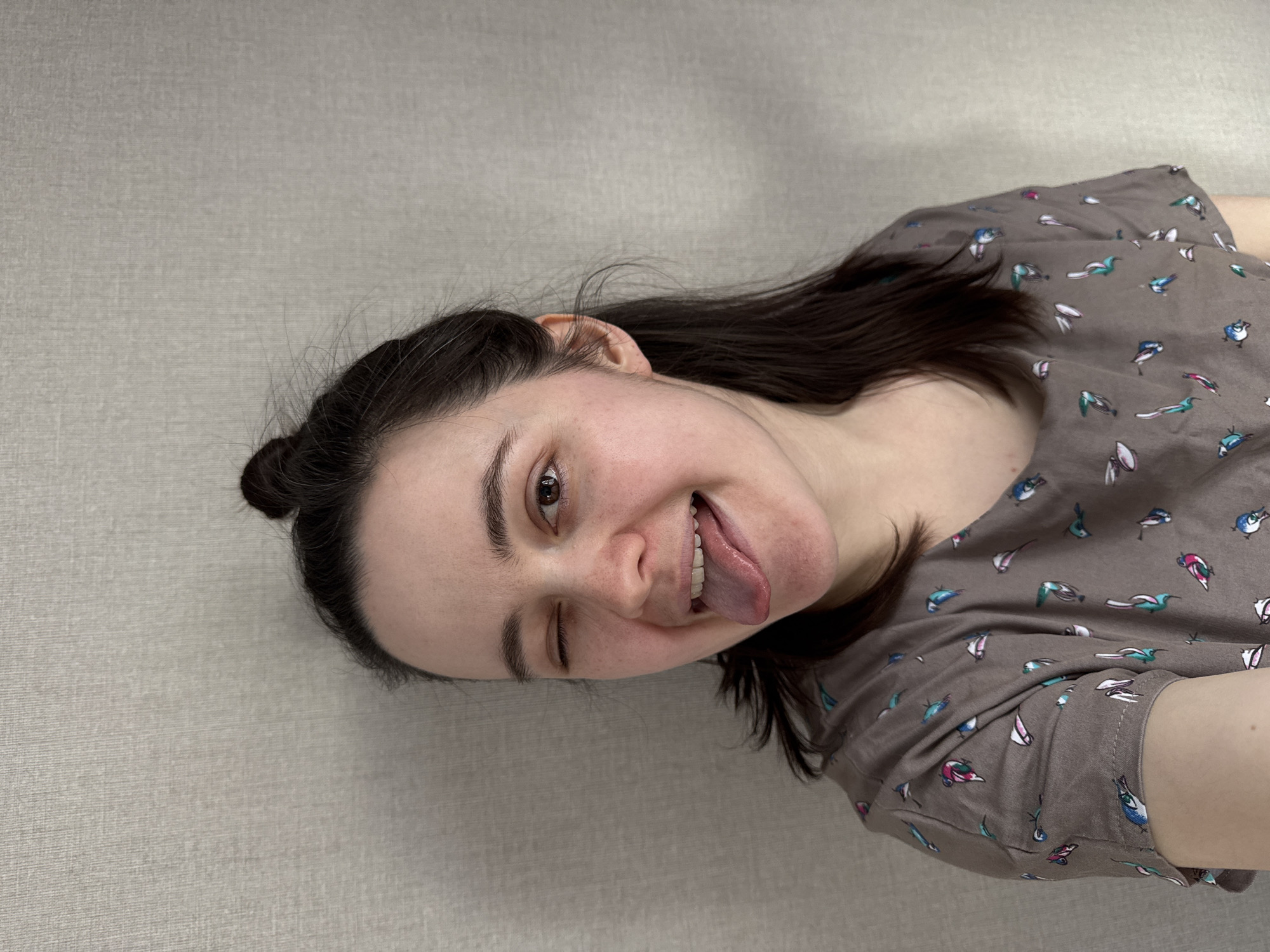 An image captures a person from the chest up, oriented diagonally with their head towards the top left and body extending to the bottom right. The individual has dark hair, partially tied back in a small bun visible on the left side of their head, with some strands loose. They are wearing a short-sleeved, greyish-brown top patterned with small, colorful birds in shades of blue, green, pink, and white.
The person is looking directly at the camera with a playful expression. They are winking with their right eye (from the viewer's perspective, their left eye) and sticking their tongue out. Their left eye (viewer's right eye) is open. Their skin is fair with visible freckles. The background is a plain, light grey textured wall, suggesting an indoor setting. There are no other visible objects or text. The overall mood conveyed is lighthearted and casual.