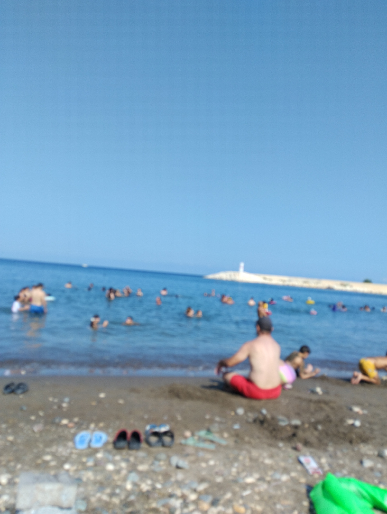 The image depicts a crowded beach scene on a clear, sunny day, likely in Erdemli, Turkey. The foreground shows a pebbly beach with several pairs of flip-flops and a bright green inflatable object. In the middle ground, a man is seated on the sand in red shorts, facing away from the camera. To his right, a child in pink shorts is playing on the sand, and further right, another person in yellow shorts is visible. The sea is filled with numerous people swimming and enjoying themselves, with a white lighthouse structure visible on a breakwater in the background. The overall atmosphere is one of leisure and recreation.