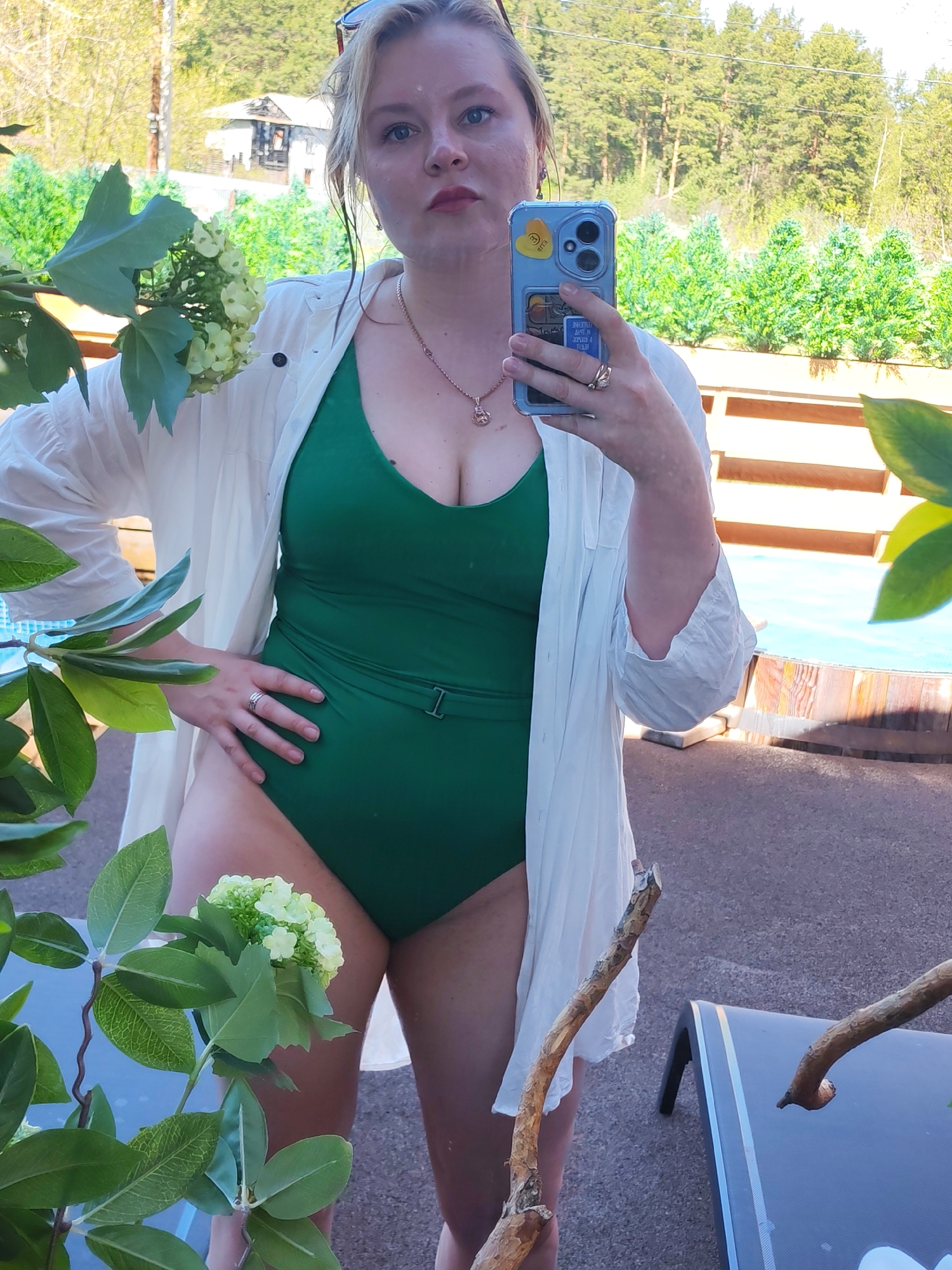A woman is taking a selfie outdoors. She is wearing a green one-piece swimsuit and a white button-down shirt. Her sunglasses are placed on top of her head. She is holding a phone with a blue case. A branch with green leaves and white flowers is in the foreground. Behind her, there is a wooden fence and greenery. A building is visible in the background, and the scene appears to be a backyard or garden setting. The location is not identifiable as Izhevsk, Russia, based on the visual information provided.
