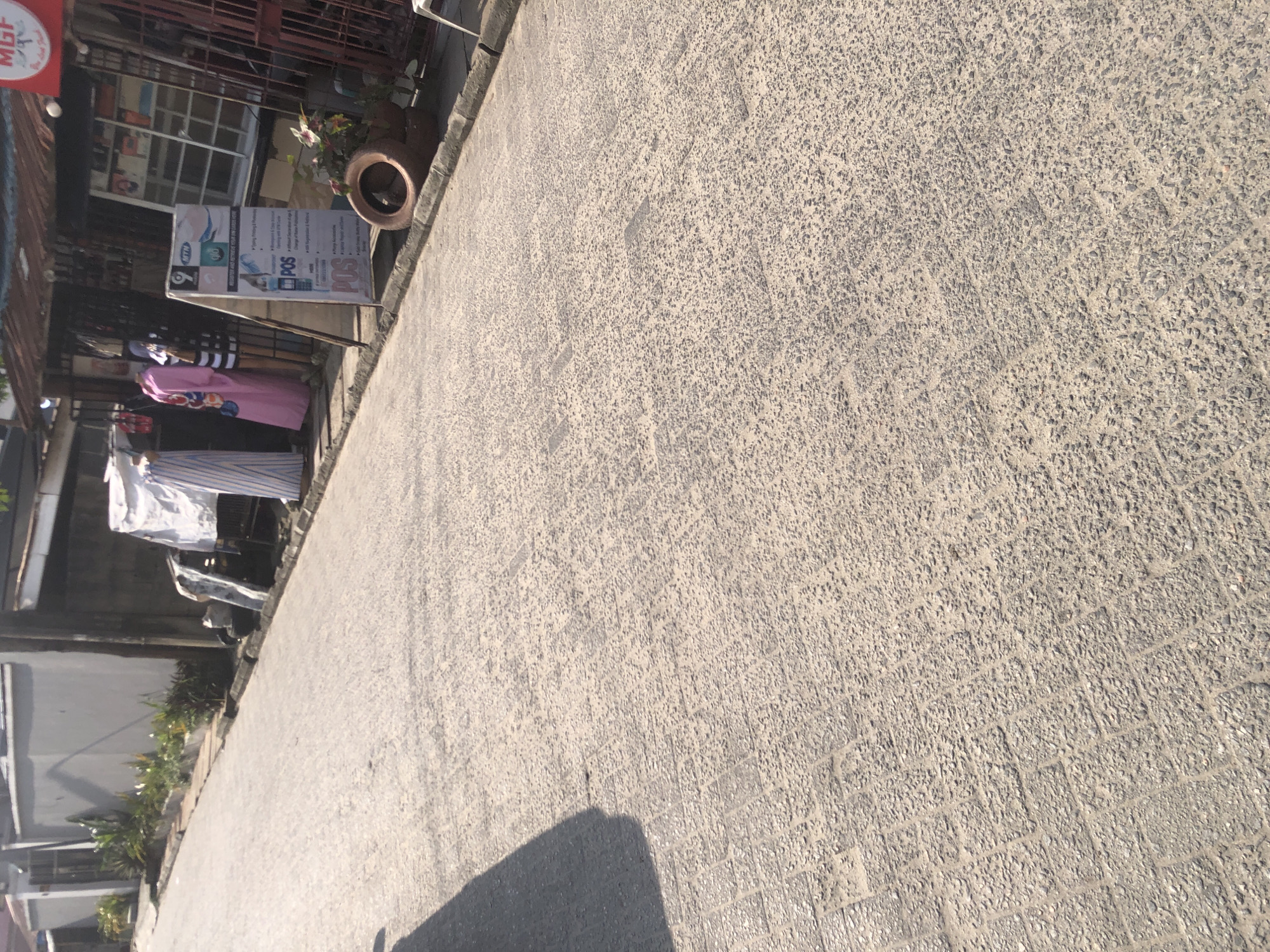 The image depicts a street scene in Ado, Nigeria, characterized by a paved road with a granular texture. To the left, a light-colored building with a dark fence is visible, bordered by greenery. On the right side of the road, a storefront displays mannequins wearing colorful clothing, and an advertising stand is positioned in front of it. The storefront appears to be a small business. The overall environment suggests a moderately populated urban area with commercial activity.