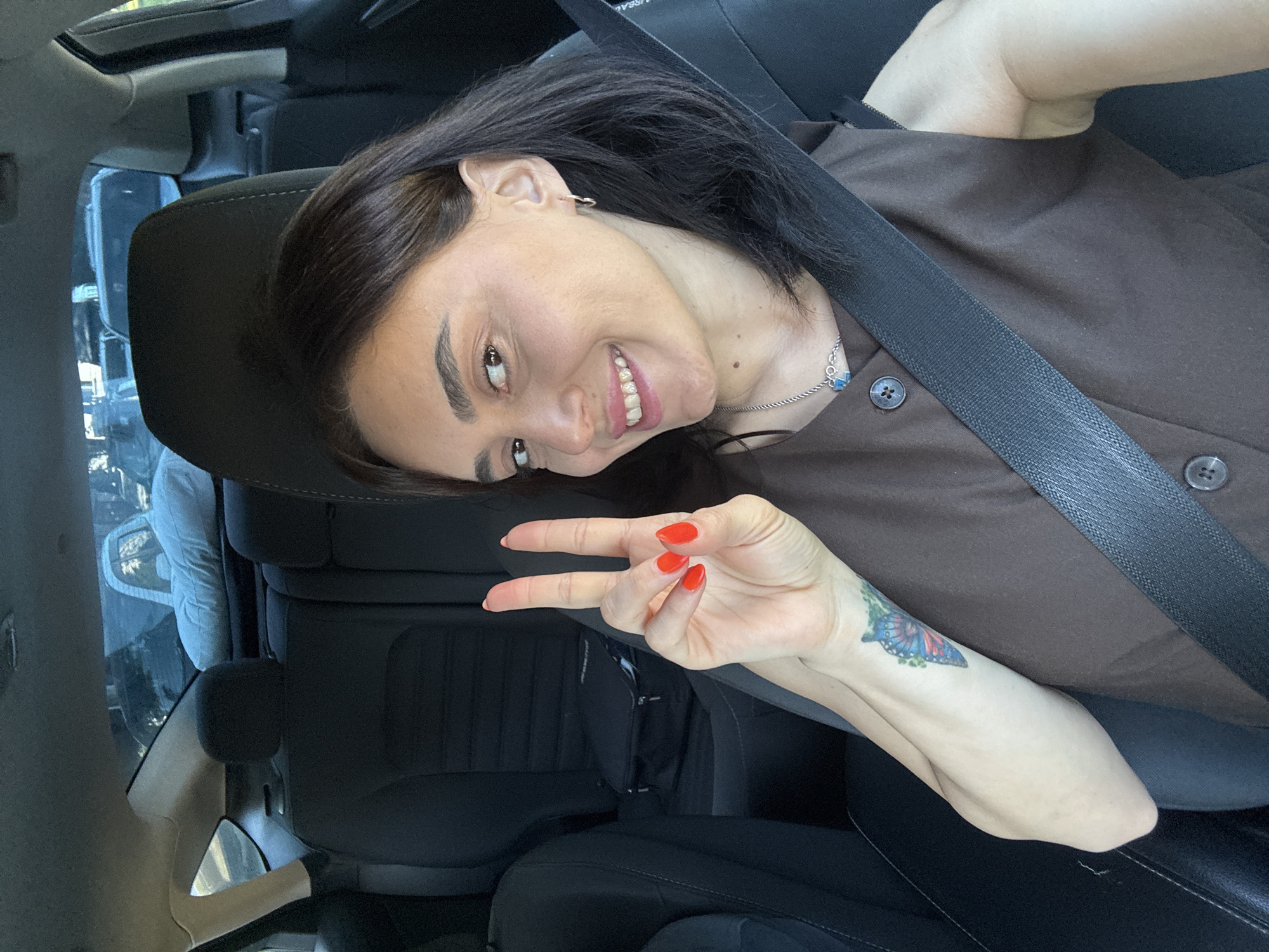 A woman with dark hair and fair skin is in the driver's seat of a car, taking a selfie. She is wearing a brown sleeveless dress with two visible buttons. Her right hand is raised, making a peace sign with bright red nail polish. A seatbelt crosses her chest. A tattoo of a butterfly is visible on her left forearm. She is smiling and looking at the camera. The background shows the interior of the car, including car seats and a pillow on the back seat. The scene appears to be during the daytime, with light filtering into the car. There is no specific indication of the location being Tbilisi, Georgia.