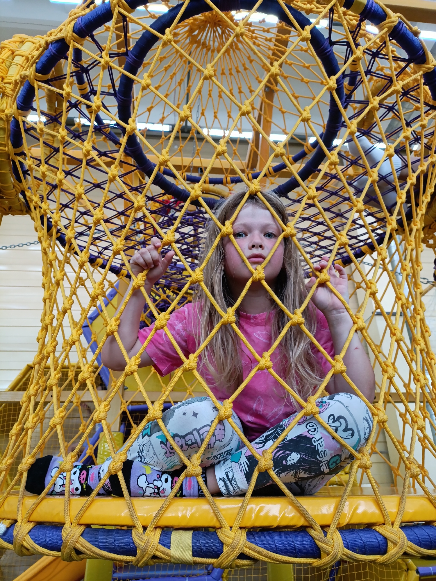 A young girl with long blonde hair is sitting inside a large, yellow rope net structure. She is wearing a pink t-shirt and patterned leggings with cartoon characters and text. She is looking towards the camera with a serious expression. Her hands are resting on the ropes of the net. The structure appears to be part of a playground or indoor play area, with other yellow and blue elements visible in the background, along with what looks like wooden paneling. The lighting suggests it is daytime, likely indoors given the bright, even illumination. The girl's pose and gaze give a sense of introspection or perhaps boredom. The text on her leggings is not legible.