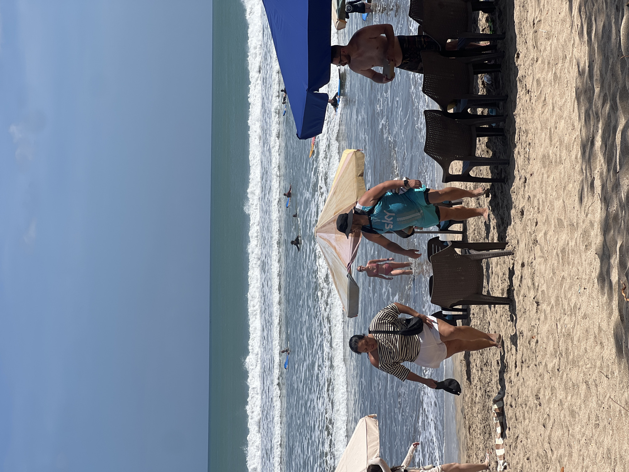 The image shows a busy beach scene on a sunny day in Kuta, Indonesia. In the foreground, there are several dark brown plastic chairs set up on the sandy beach, some partially in the shallow water. Two beach umbrellas, one large blue and one smaller tan and yellow striped, provide shade. A woman in a striped shirt and white shorts is walking away from the water, holding a black hat in her left hand. To her right, a man wearing a turquoise tank top with "sky" printed on it and black shorts is also walking on the beach. Further in the background, a man with glasses and patterned swim trunks is standing in the shallow water. A woman in a coral-colored bikini is walking in the water. Beyond them, several people are visible in the ocean, some on surfboards and others swimming. The waves are moderate, and the sky is a clear blue.