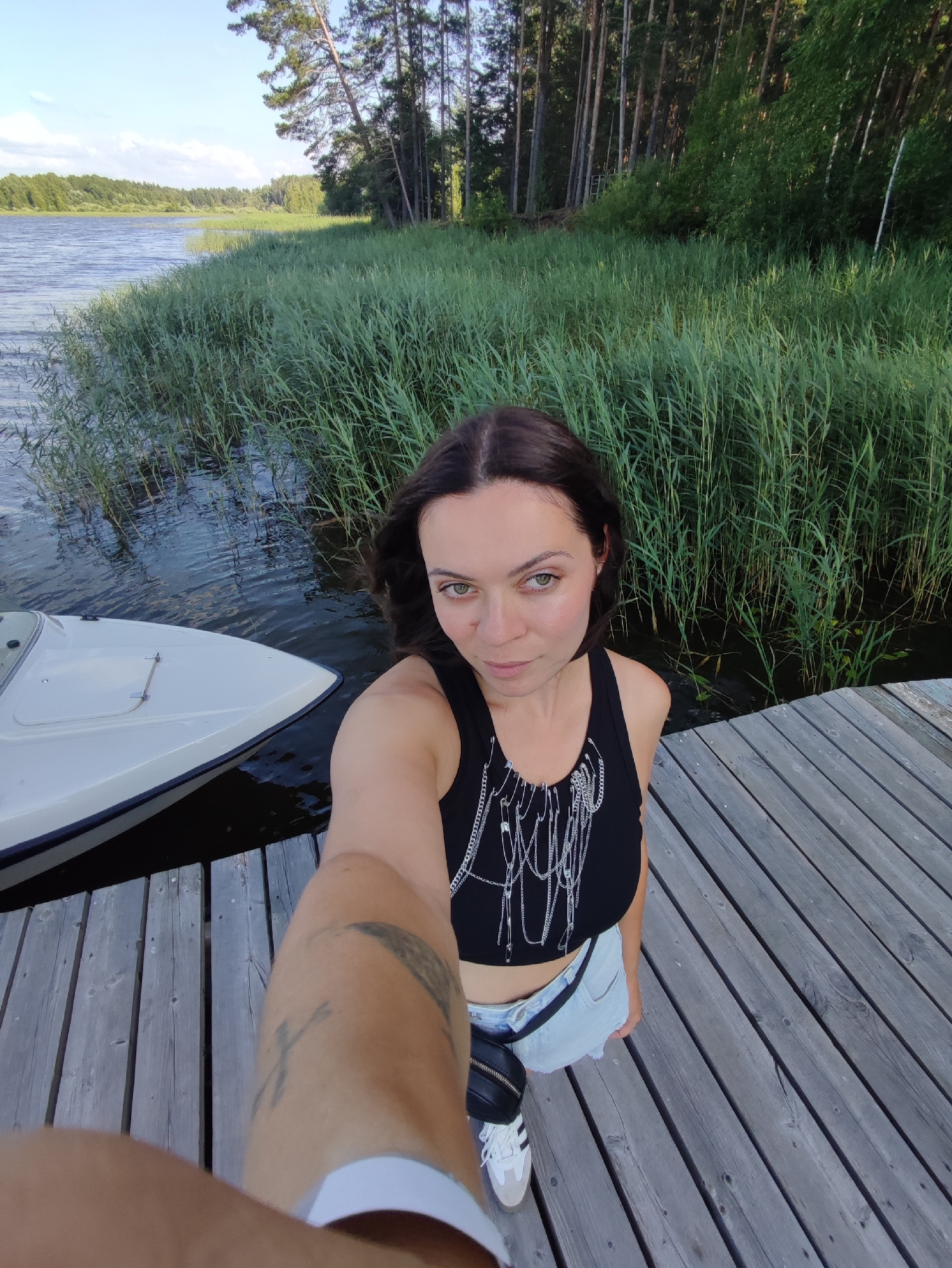 A woman with brown hair takes a selfie on a wooden pier. She is wearing a black crop top with a chain necklace and light blue denim shorts. She has white sneakers and a black fanny pack. To her left, a white boat is docked. The pier is next to a body of water with tall green reeds. In the background, there is a forest with tall trees. The sky is blue with some clouds. The image does not depict any recognizable landmarks or features specific to the city of Chaykovsky, Russia.