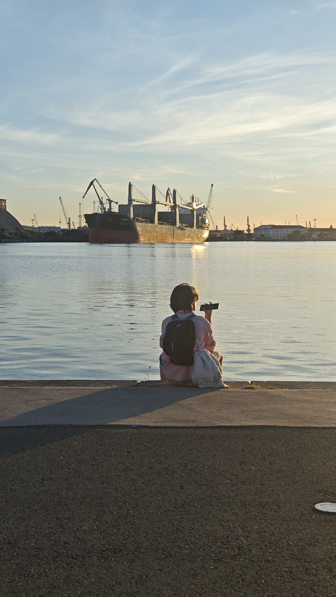 A woman sits on a concrete embankment in Burgas, Bulgaria, facing a body of water and a large cargo ship docked in the distance. The woman is wearing a pink shirt and has a dark-colored backpack on her back. She holds a smartphone in her hand, possibly taking a photograph. The sky is a mix of blue and orange hues, suggesting it is either sunrise or sunset. In the background, several cranes and buildings are visible along the waterfront. The foreground consists of a paved area, with a discernible shadow cast by the embankment.