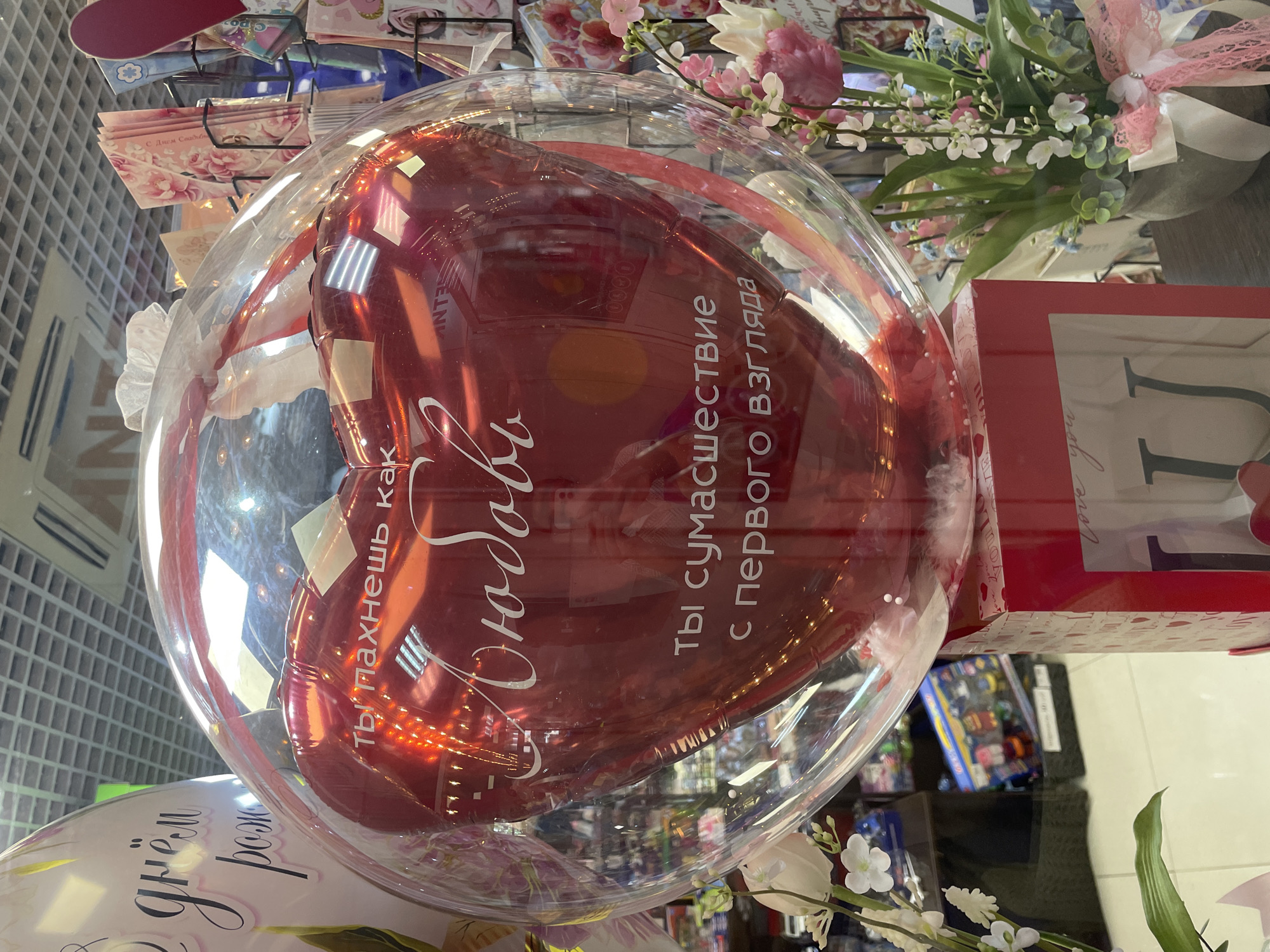The image shows a balloon in the shape of a red heart inside a clear plastic sphere. The balloon has white text written on it in Russian, which reads "ты пахнешь как Любовь" and "ты сумасшествие с первого взгляда". This translates to "you smell like Love" and "you are madness from the first glance". The balloon is sitting on top of a red box, which is decorated with hearts and the word "love". In the background, there are shelves filled with greeting cards and other gifts, suggesting a retail environment, possibly a gift shop or flower shop. The lighting suggests an indoor setting with overhead fluorescent lights. The overall scene conveys a romantic theme. The location information provided is that the scene is in the city of Sochi, Russia. However, there are no visual cues within the image that specifically confirm this location.