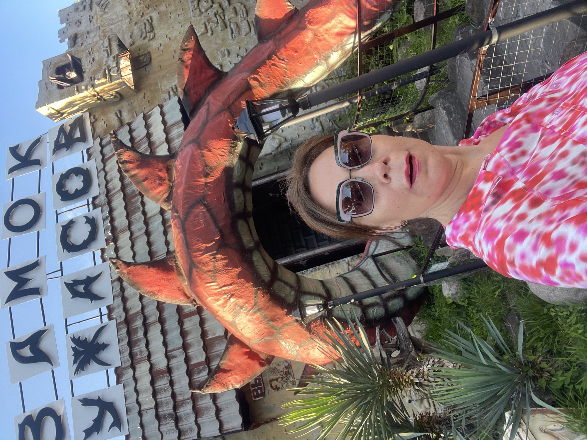 A woman wearing sunglasses and a pink and white tie-dye shirt takes a selfie in front of a building with a large red dragon-like structure over the entrance. The building has the words "ЗАМОК УЖАСОВ" (Castle of Horrors) written in large black letters above the entrance. The building's facade is textured and appears to be made of stone or concrete, with a tiled roof over the entrance. To the left of the entrance, there is a yucca plant. The scene is likely located at an amusement park or attraction in Sochi, Russia, given the signage and thematic design. The woman is positioned in the foreground, with her face and upper body visible. The lighting suggests it is daytime.