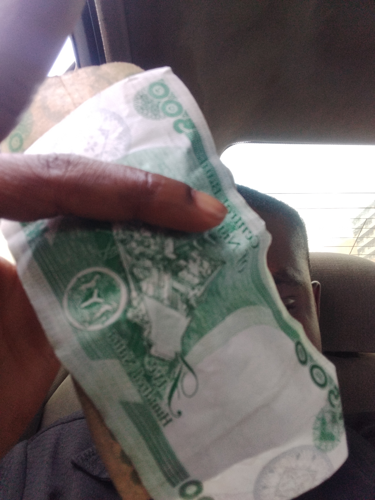 A person's hand, with dark skin tone, holds up a Nigerian 200 Naira banknote. The banknote is predominantly white with green lettering and designs. The number "200" is visible in green circles on the right side of the note, and a stylized symbol with three stars within a circle is on the left. The person's thumb and index finger are wrapped around the note, obscuring part of it. The background is blurred, suggesting the photo was taken inside a vehicle. The head and shoulder of another person are visible to the right of the banknote. The scene appears to be indoors, possibly a car interior, with a bright light source visible in the upper left corner, hinting at daytime. The image is a close-up of the banknote and the hand holding it, with the surrounding environment out of focus.