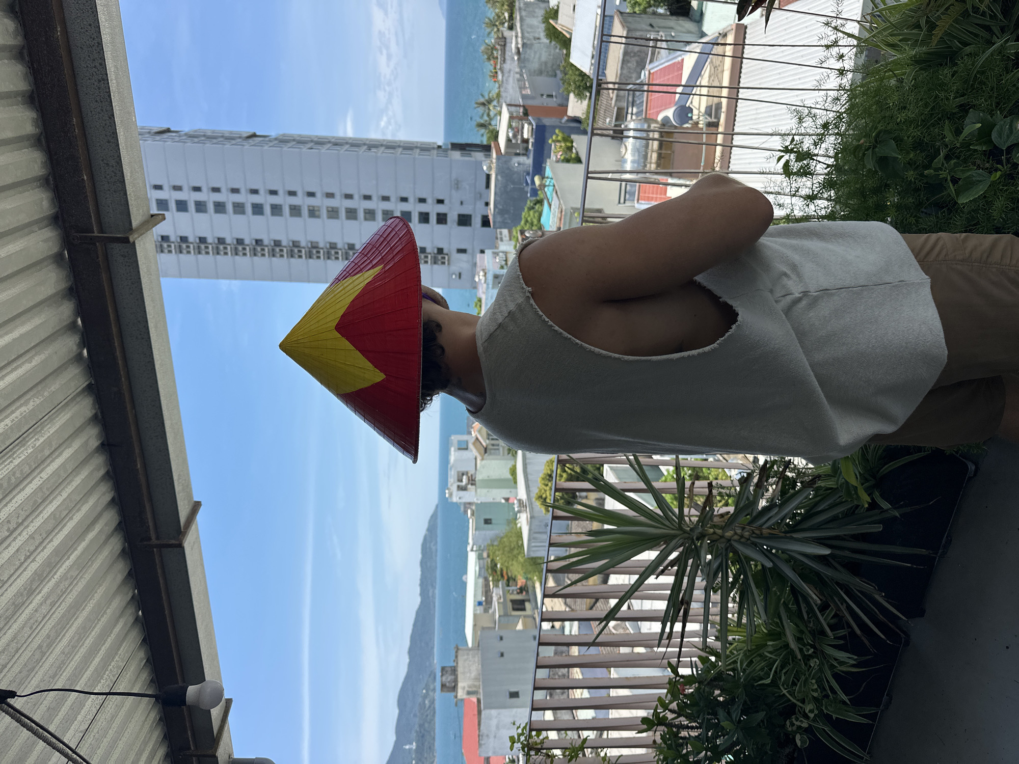 A person wearing a red and yellow conical hat stands on a balcony overlooking a coastal city. The person is viewed from the back, wearing a light-colored sleeveless shirt and tan shorts. The balcony is furnished with planters containing various green plants, including a spiky yucca-like plant. In the background, a dense urban landscape of buildings is visible, extending towards the ocean. A tall, modern skyscraper stands prominently in the middle ground, with other residential buildings of varying heights and colors scattered throughout. The sea is visible beyond the city, with distant mountains forming the horizon under a clear blue sky. The scene appears to be in Đà Nẵng, Vietnam, given the characteristic architecture and coastal setting.