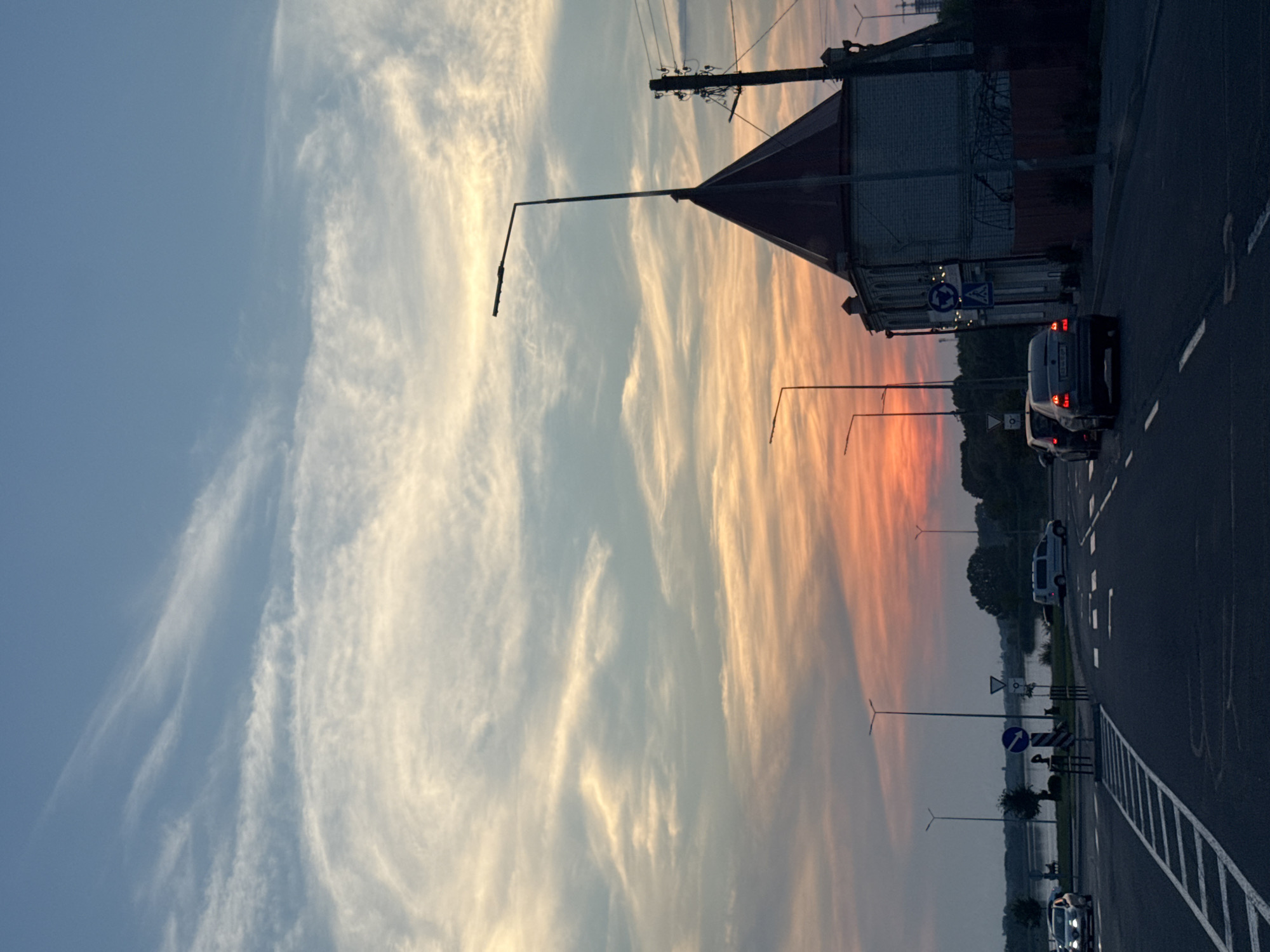 The image depicts a road scene at sunset in Vinnytsia, Ukraine. The sky is a prominent feature, displaying a gradient of colors from light blue to vibrant orange and pink hues, indicative of a dramatic sunset. Wispy clouds streak across the sky, catching the fading sunlight.
On the right side of the image, a paved road runs horizontally, bordered by a dashed white line marking. Several vehicles are present on the road, including a dark-colored car with its taillights illuminated, suggesting it's either braking or its lights are on. Further down the road, another vehicle, possibly a white van, is visible.
Along the roadside, lampposts stand tall, some with their lights on. A building with a distinctive triangular roof is visible in the background, with power lines running overhead. Road signs are also present, including a circular sign with a blue background and a white arrow indicating a roundabout, and another sign with a black border and a white symbol, possibly a warning sign.
The scene appears tranquil, with the vibrant sunset lending a peaceful or perhaps dramatic atmosphere. The presence of the road, vehicles, and buildings suggests an urban or semi-urban setting. The time of day is clearly sunset, and the weather appears clear with the exception of the clouds. No people are clearly identifiable as subjects in the image, although figures may be implied within the vehicles or distant scenery.