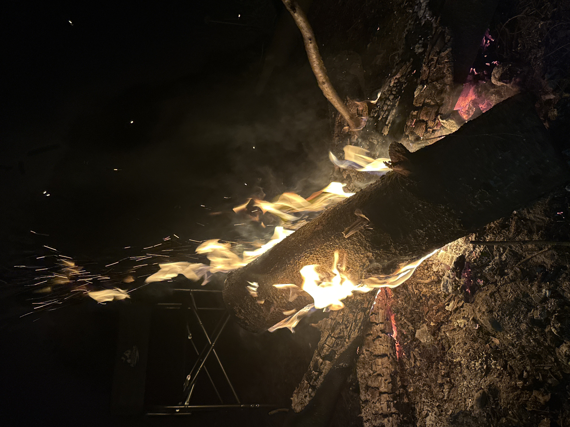 The image depicts a campfire at night in Юзуфоўскі сельскі Савет, Belarus. A large log is burning with visible flames, sparks, and smoke. The fire is surrounded by other charred wood and ash. A metal stand is visible on the left side of the frame.