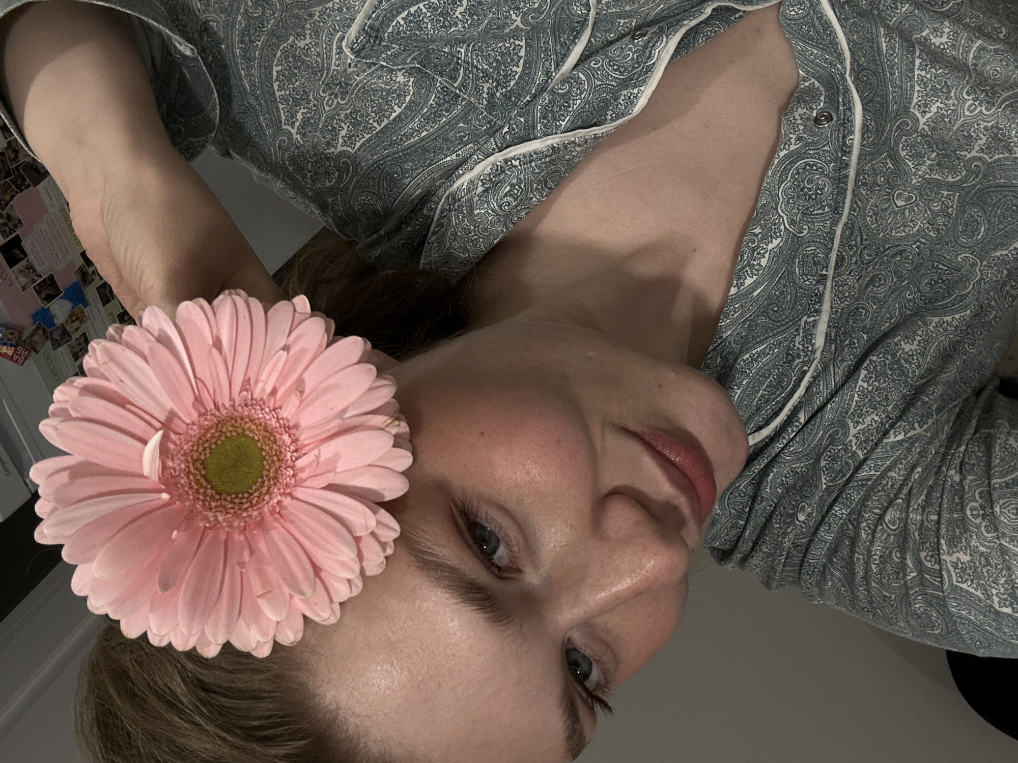 A light-skinned female with light hair is positioned at an angle to the camera, wearing a button-down shirt with a paisley print. She holds a pink gerbera daisy near her face. The background is out of focus, but appears to be an interior setting with a wall partially visible. The location in the city of Kazan, Russia, cannot be verified from this image.