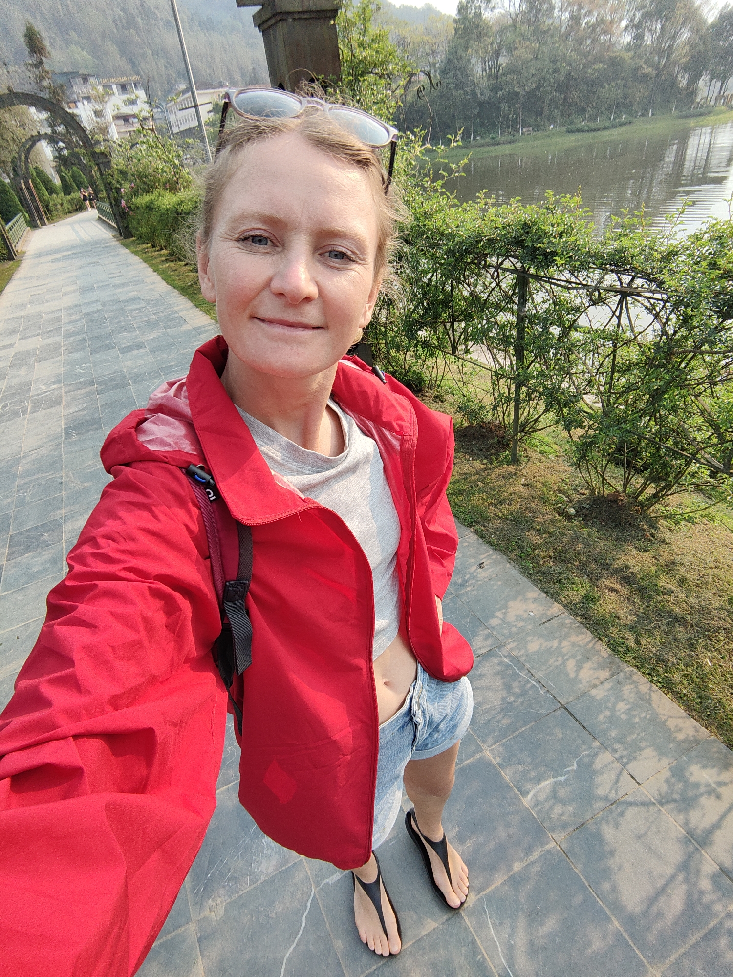 An adult fair-skinned female is depicted in a selfie, standing outdoors on a grey tiled pathway in the city of Sa Pa, Vietnam. She is smiling faintly and directly facing the camera. Her light-colored hair is pulled back, and she wears sunglasses perched on her head. Her attire consists of a bright red unzipped hooded jacket over a light grey short-sleeved t-shirt, light blue denim shorts, and black minimalist sandals. A black strap, consistent with a backpack or bag, is visible over her right shoulder.
To her immediate right, a row of green bushes and a dark metal fence border a body of water, likely a lake, which reflects distant trees under a hazy sky. To her left, the paved path continues past a series of dark metal arched trellises and further greenery. In the distant background, white buildings are situated on a forested hillside beneath hazy mountains. The scene is illuminated by natural light, casting subtle shadows on the pathway.