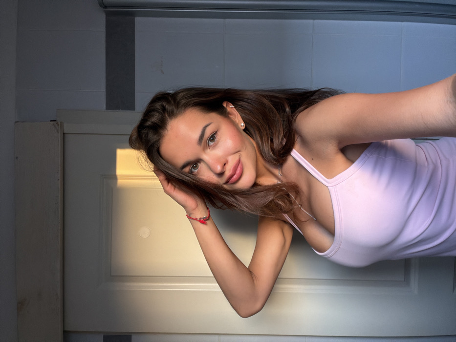 An indoor, close-up portrait depicts a woman in her late 20s or early 30s, positioned against a light-colored background. The scene is depicted indoors in Tel-Aviv, Israel. She possesses medium-brown hair, styled with volume, and a fair complexion. Her facial features include full lips, pronounced cheekbones, and dark eyes. She is wearing a light pink, spaghetti-strap tank top. Her right arm is raised, with her hand gently touching the side of her head, affecting a pose. A small, round stud earring is visible in her right ear, and a thin silver-colored necklace with a small pendant hangs around her neck. A red string bracelet with an attached charm is present on her right wrist. Her expression is a slight smile, and she is looking directly into the camera lens.
The background on the left consists of a light-colored, possibly white, panel door, partially illuminated by a warm, golden light source entering from the upper left, casting a distinct patch of sunlight. Above the door, a horizontal light-colored wooden beam or trim is visible. The background on the right features a wall composed of light gray or muted bluish-gray rectangular tiles. A vertical chrome or metallic bar, consistent with a shower enclosure frame, is visible along the far right edge of the frame. The lighting suggests ambient interior illumination mixed with direct sunlight from an external source.