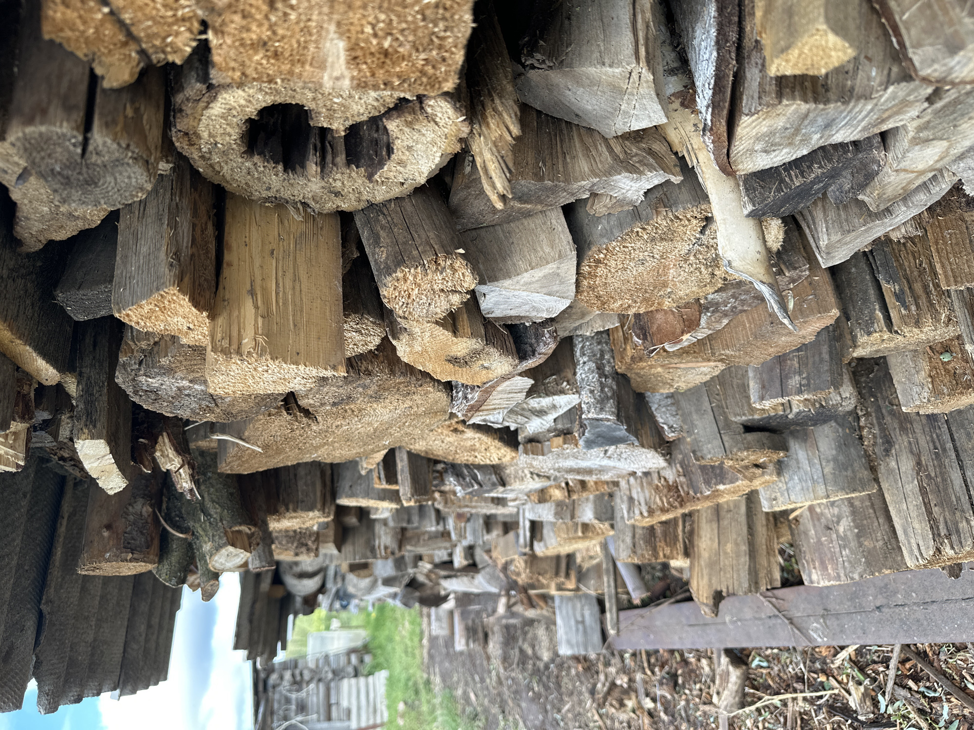 The image depicts a close-up view of a stack of firewood located outdoors, likely in Среднее Кечево, Russia, given the prompt's instruction. The firewood consists of roughly cut pieces of wood, varying in size and shape, stacked haphazardly. Some pieces appear to be bark-covered, while others are cleanly split. The wood is of various colors, ranging from light brown to gray. The stack's orientation gives the impression of looking upwards at the woodpile. In the background, a glimpse of a blurred landscape can be seen, suggesting the woodpile is located in an open area near a garden or yard.