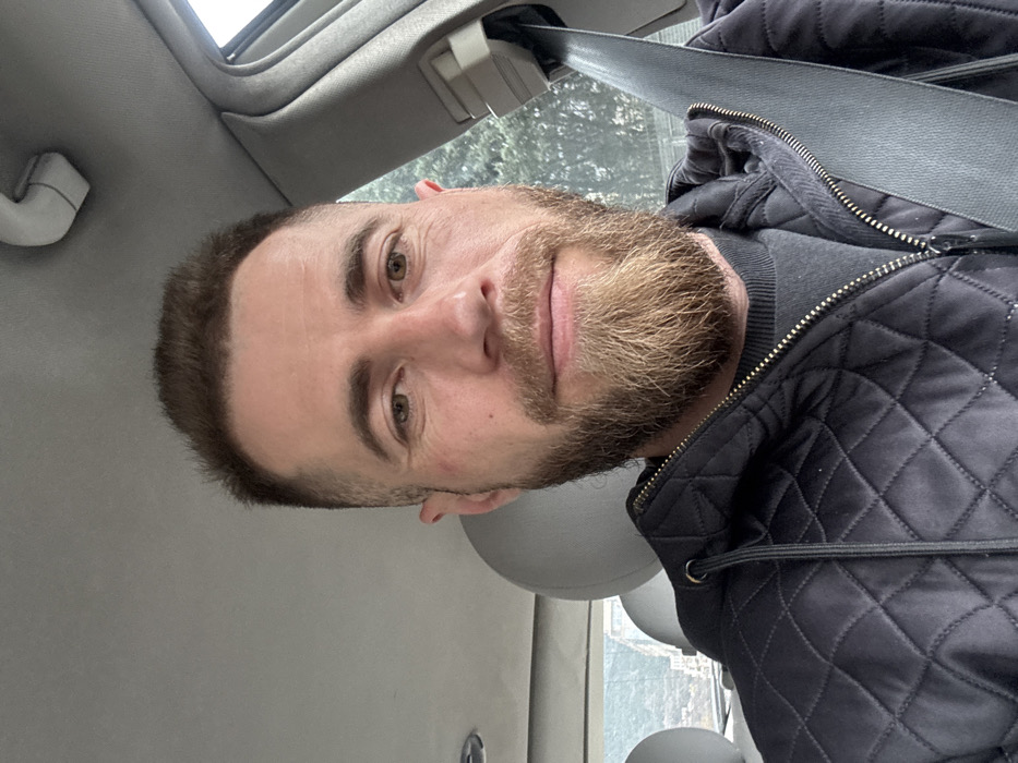 An adult male is seated inside a vehicle, captured in a selfie-style shot from the chest up. He has short, dark brown hair with faded sides and a full, light brown to blonde beard and mustache. His eyes are light brown. He wears a black quilted jacket over a dark grey crew-neck sweater and is secured by a black seatbelt extending diagonally across his chest and right shoulder. The vehicle's interior features a light grey headliner and two light grey headrests are visible in the background. Through the rear left passenger window, a mountainous landscape with scattered white or light-colored buildings on a hillside is visible, consistent with the topography of Budva, Montenegro. Through the right window, blurred green foliage is discernible. The man maintains a neutral facial expression.