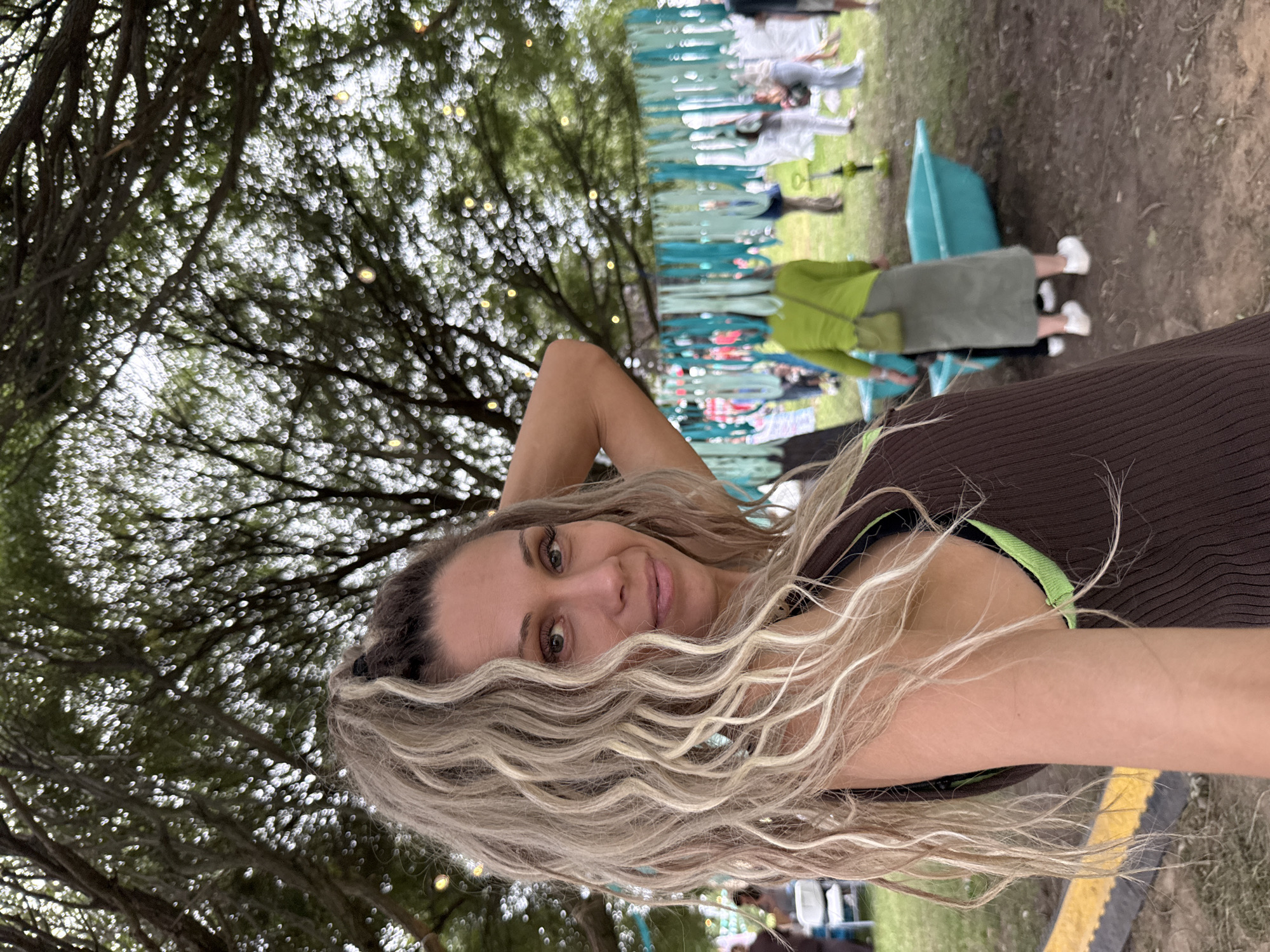 An outdoor, eye-level, medium shot shows a woman in the foreground with her right hand raised to her head. She has blonde, wavy hair that is pulled back into a high ponytail, and she is wearing a brown tank top with green trim. The woman is looking at the camera with a slight smile. In the background, there are trees with bokeh lights, and a group of people are visible. There is also a teal bathtub in the middle ground. The ground is covered in dirt and grass. The scene appears to be at a festival or outdoor event.