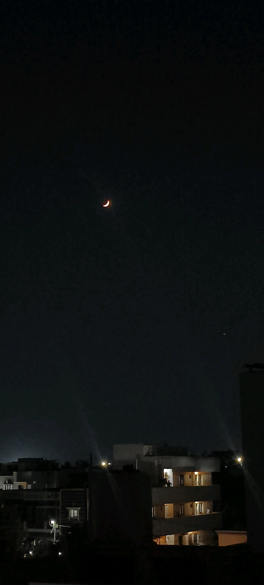 The image shows a night-time view of the city of Kundrathur, India. In the dark sky, a crescent moon is visible, as well as a small, bright star. Below the sky, the silhouette of buildings and rooftops is apparent. Some buildings have lights on, indicating occupancy. A few of the structures appear to be residential or commercial buildings, with balconies and windows visible. The overall scene depicts a typical urban environment at night.