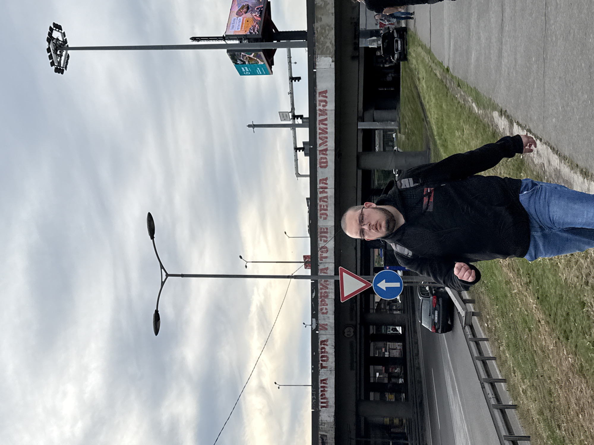 A man stands beside a highway overpass in Vozdovac Urban Municipality, Serbia. Graffiti is visible on the overpass's support structure. A road sign directing traffic to turn left is next to the man. Cars are visible on the highway. Streetlights and billboard are in the background. The sky is overcast.