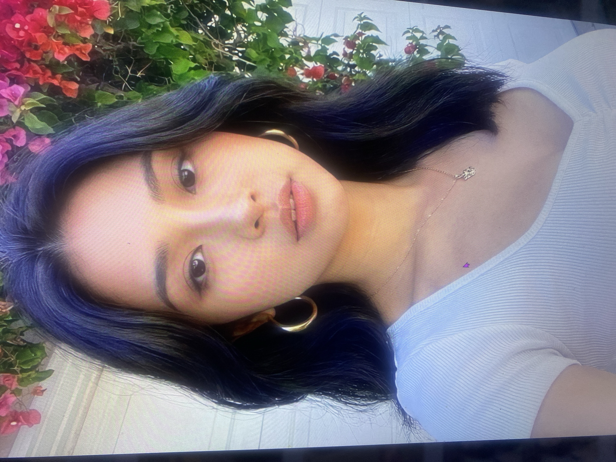 A young Asian woman with dark, wavy hair is captured in a close-up portrait. She is wearing a white ribbed V-neck top and a delicate gold necklace with a small pendant. Large gold hoop earrings adorn her ears. Her makeup includes winged eyeliner and lip gloss. The background features a wall, possibly part of a building, and vibrant pink and red bougainvillea flowers. The lighting suggests an outdoor setting during the daytime. The image does not provide any information related to the city of Samara, Russia.