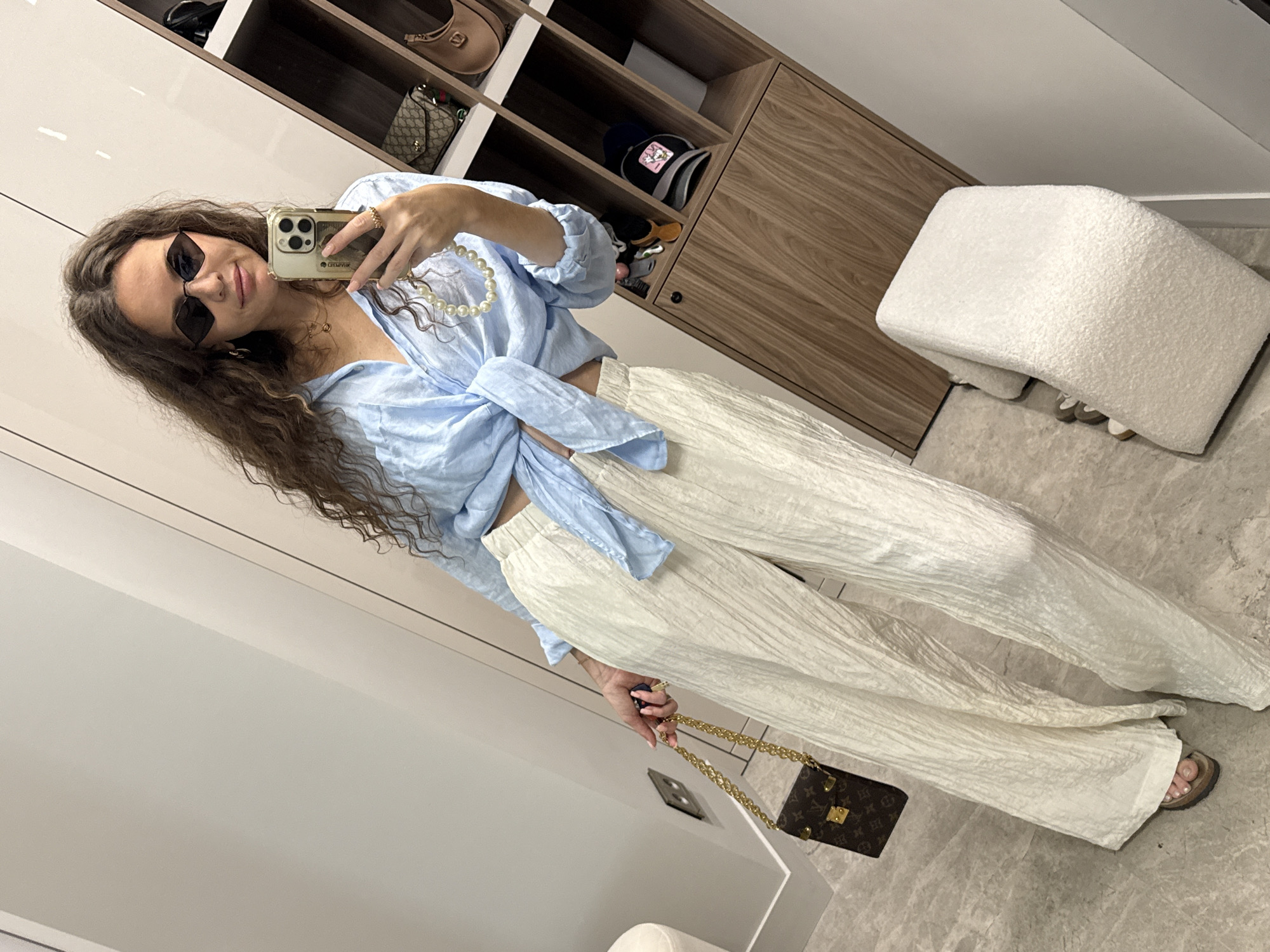 A woman with long, curly brown hair is taking a selfie in what appears to be a dressing room or closet area. She is wearing a light blue, cropped, tie-front blouse and loose, cream-colored, crinkled pants. She accessorizes with black, diamond-shaped sunglasses, a pearl necklace, gold hoop earrings, and a small brown designer handbag with a gold chain strap. She is holding a smartphone in her right hand, capturing the reflection in a mirror. The background reveals a wooden shelving unit filled with bags and accessories, and a white upholstered bench. The floor is a light gray marble tile. There is no discernible indication of the city being Moscow, Russia.