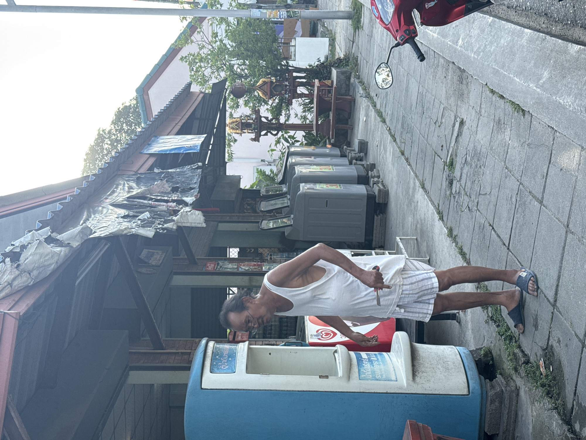 A man wearing a white tank top and striped shorts stands on a sidewalk in front of a blue vending machine. He is holding a small knife in his right hand and appears to be interacting with the machine. Several washing machines are lined up behind him. In the background, there are buildings and trees. A red motorcycle is parked on the right side of the image. The scene is located in the city of Ko Samui District, Thailand.