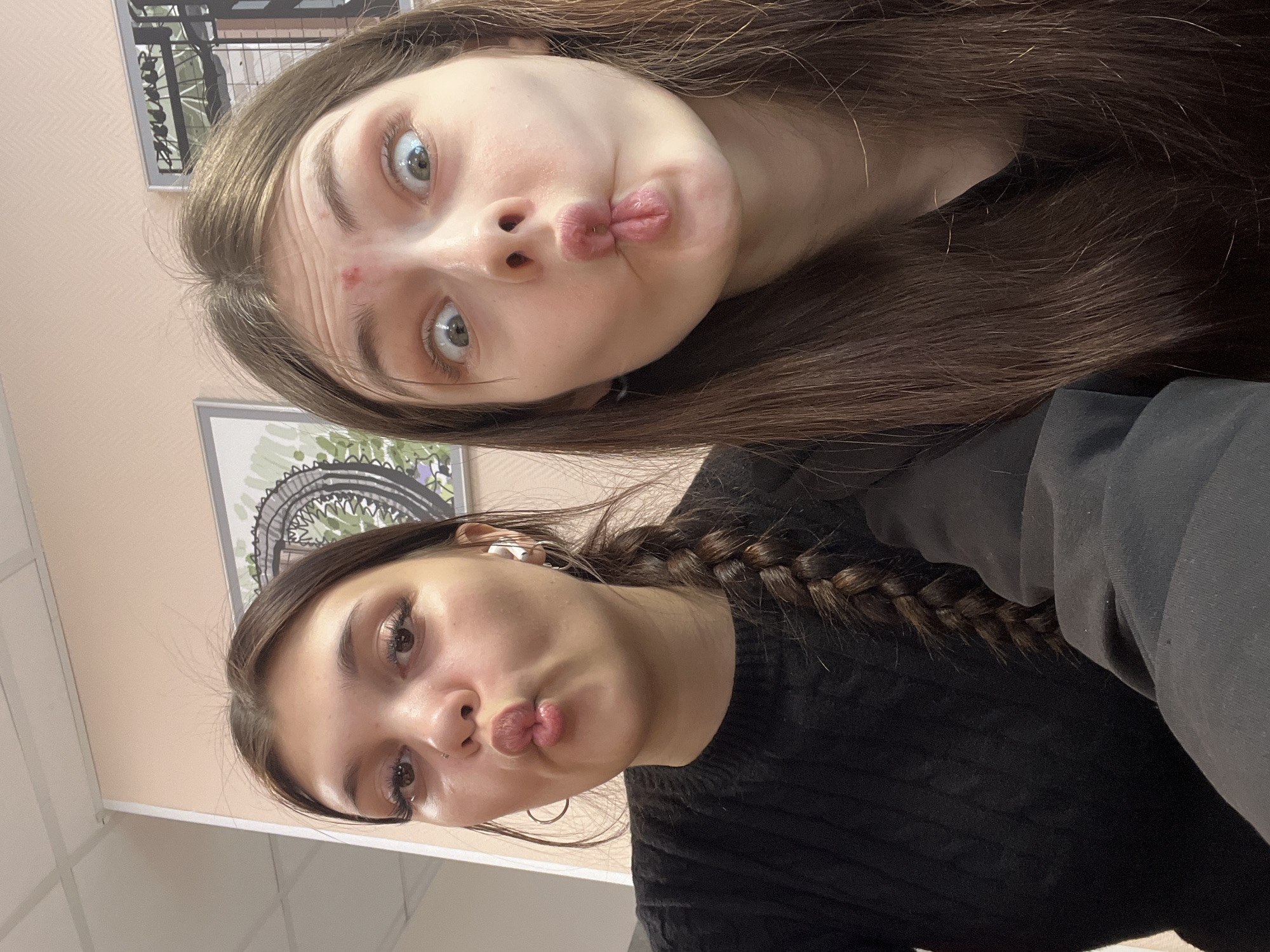 This media file is a selfie-style photograph featuring two young women. The image is oriented vertically, appearing as if it was originally taken horizontally and then rotated 90 degrees clockwise, causing the subjects to appear to be looking upwards. The setting is indoors, likely in Saint Petersburg, Russia, with plain, light-colored walls and visible white ceiling tiles at the bottom of the frame.
The woman positioned in the upper half of the image has long, straight light brown hair and prominent blue-grey eyes. She has a small red blemish on her forehead and is making a "duck face" or pouty expression with her lips.
Below her is the second woman, who has dark brown hair styled in a braid that rests over her shoulder. She has brown eyes and is also making a similar puckered-lips expression. She is wearing a black knitted sweater and has hoop earrings, with an earbud visible in her left ear.
In the background, two framed artworks are visible on the wall, showing abstract or stylized illustrations, possibly of landscapes or architectural elements. The overall activity is a casual and playful moment captured between the two women. Their expressions convey a lighthearted and friendly mood. There is no visible text in the image. The lighting suggests an indoor environment, likely during the daytime.