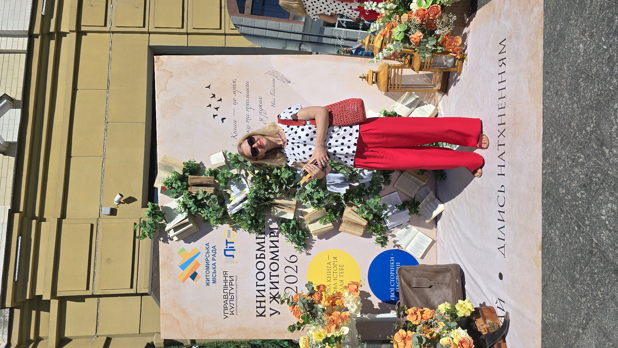 The image shows a woman standing in front of a large banner that appears to be part of an outdoor event in Zhytomyr, Ukraine. The banner has text in Ukrainian, including "ЖИТОМИРСЬКА МІСЬКА РАДА" (Zhytomyr City Council) and "КНИГООБМІН У ЖИТОМИРІ 2026" (Book Exchange in Zhytomyr 2026). The banner is decorated with artificial greenery and open books, creating a tree-like effect. The woman is wearing a black and white polka dot top and red pants, and she is holding two books. To her left, there are floral arrangements and a vintage suitcase. To her right, there are decorative lanterns. In the background, the facade of a building is visible. The ground in front of the banner has text that reads "ПОЧАЙ - ДІЛИСЬ НАХТХЕННЯМ" (Listen - Share Inspiration). The overall scene suggests a cultural event related to books and reading.