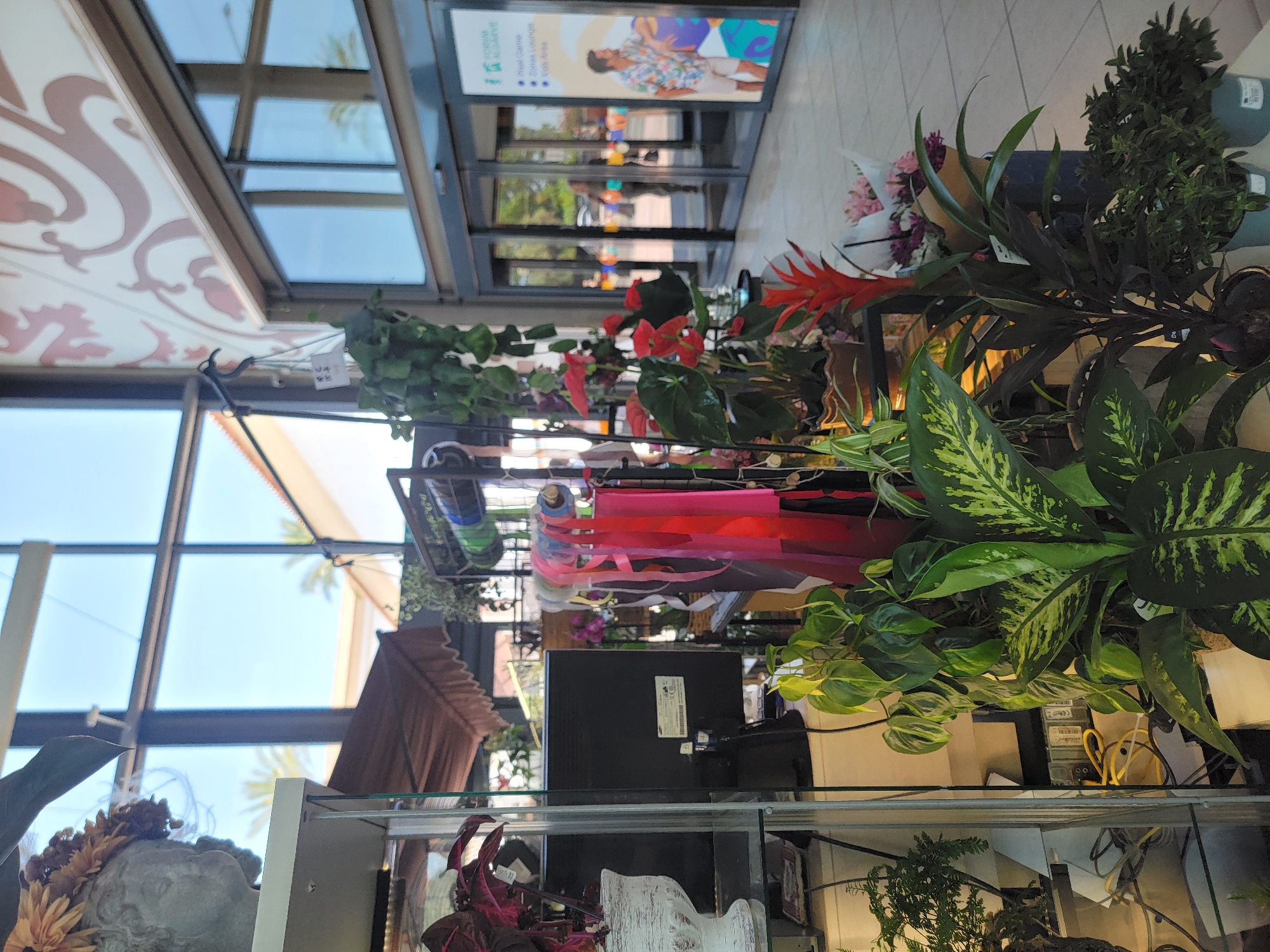This image depicts an indoor plant display situated in a bright, modern commercial space, possibly a shopping center or mall, with large glass windows and doors leading outside. The display features a variety of plants, including leafy green varieties and flowering plants with red and pink blooms. Rolls of colorful ribbon, likely for gift wrapping or floral arrangements, are also visible. In the background, glass doors reveal the exterior, where palm trees and a clear blue sky suggest a sunny, warm climate. A large sign on the glass doors displays text in English and Portuguese, along with graphics of people and icons, advertising amenities such as "Pixel Game," "Zonas Lounge," and "Kids Area," which are likely services offered at this location. The presence of the sign, which features "BOM DIA ALGARVE" and mentions "Faro," strongly indicates that this scene is located in the city of Faro, Portugal, within the Algarve region. There are no individuals prominently featured in the foreground, but several blurred figures can be seen through the glass doors in the background. The overall impression is one of a vibrant and welcoming retail environment.