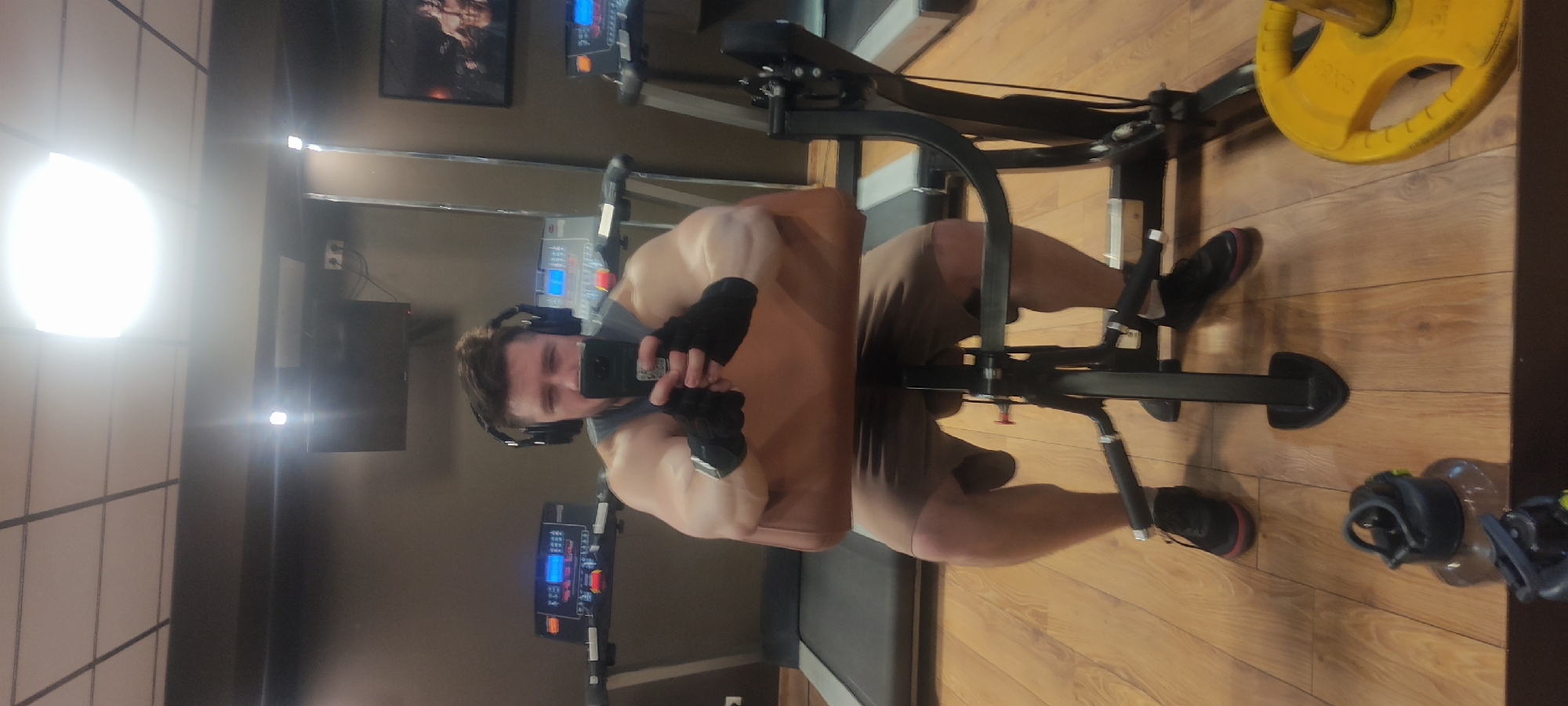 A man is taking a selfie in a gym. He is wearing headphones and gloves. He appears to be using a leg extension machine with a yellow weight plate. In the background, there is a treadmill and a monitor. The scene appears to be indoors with wood flooring and tiled walls. Without additional data, the specific location in Fryazino, Russia, cannot be confirmed.