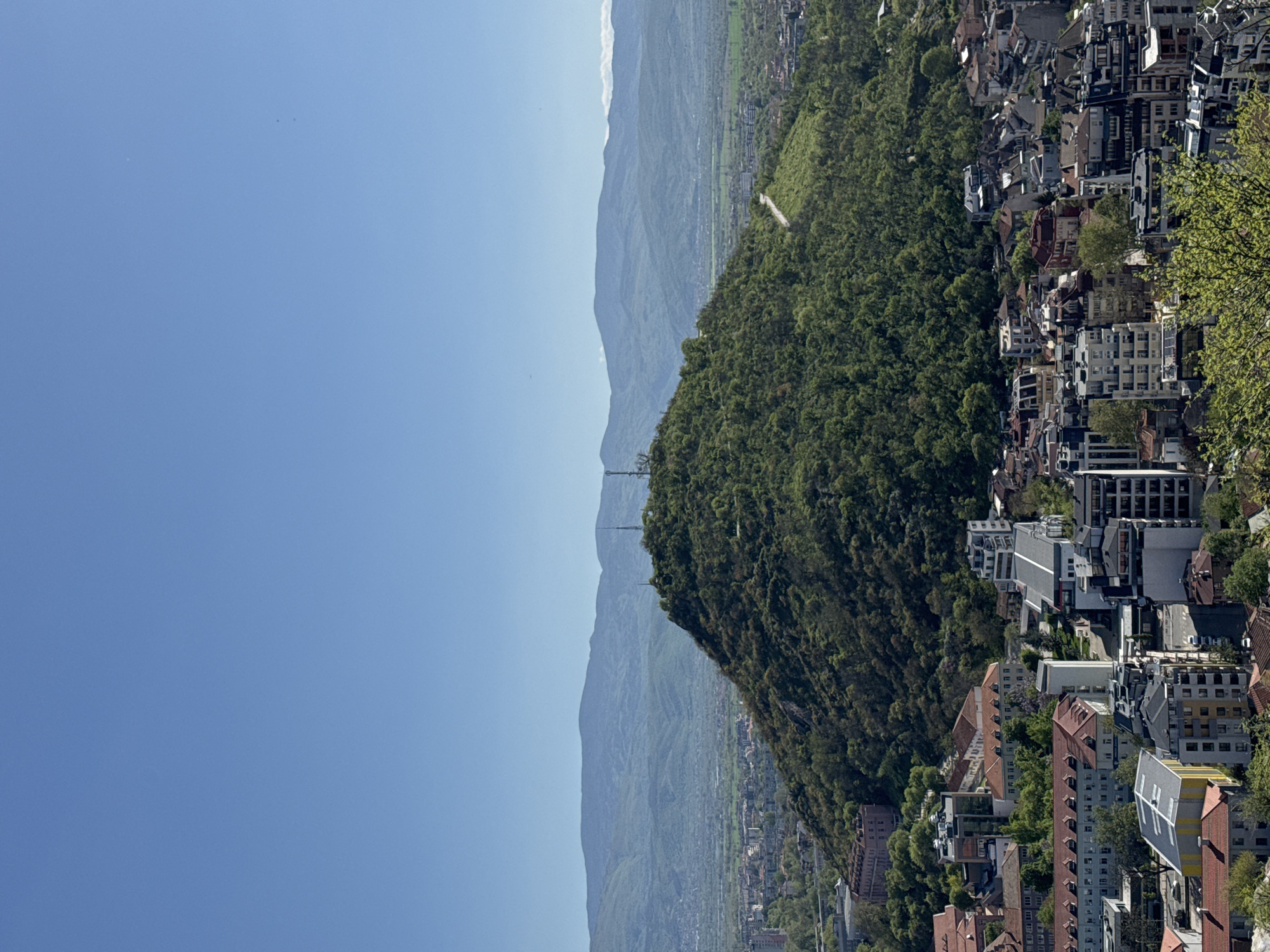 The image shows a view of Plovdiv, Bulgaria, oriented vertically with the sky on the left. Mountains are visible in the background, partially obscured by haze. A forested hill occupies the center of the frame, sloping down towards a densely built-up urban area to the right. Buildings are of varying heights and architectural styles, including both residential and commercial structures. Trees are interspersed among the buildings, adding green patches to the urban landscape. A transmission tower is visible on top of a mountain. The sky is clear blue, suggesting good weather conditions.