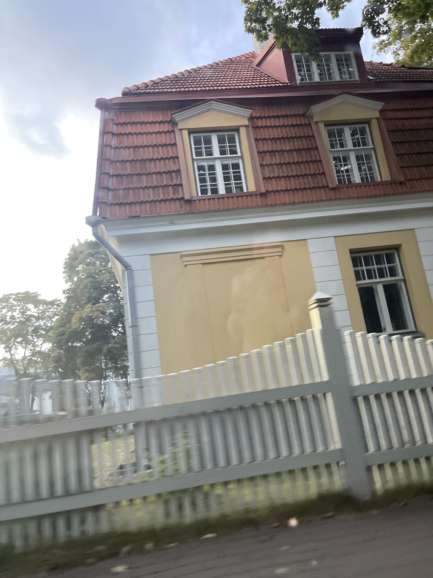 The image shows a building with a red tile roof and yellow walls, located in Tallinn, Estonia. The building has several windows with white frames and grid panes. A white picket fence is in the foreground, obscuring parts of the lower section of the building and the surrounding vegetation. Trees with green leaves are visible behind the fence and to the left of the building. The sky above is overcast.