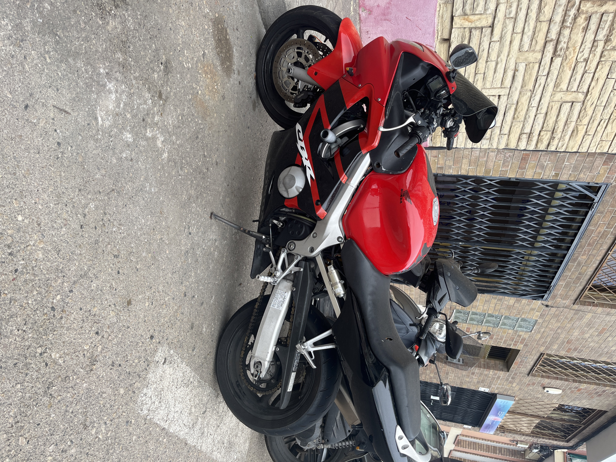 A red and black Honda CBR sport motorcycle is parked on a grey concrete street in front of a brick building with metal security gates. The motorcycle is the primary subject, positioned at a slight angle. A dark-colored scooter is parked behind it, partially obscured. The building features a rough-textured stone facade on the left and red brick on the right. The street is marked with white lines. The scene appears to be an outdoor urban environment.