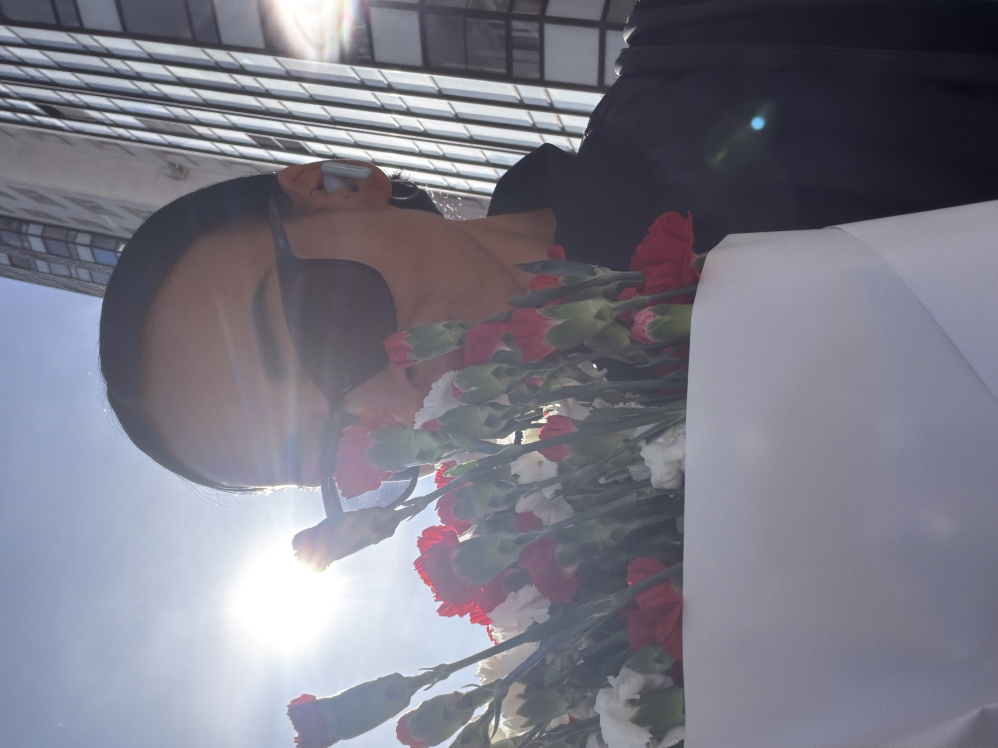 The image features a person holding a bouquet of red and white carnations. The person has dark hair and is wearing sunglasses. Behind the person, a tall building with a grid-like facade is visible. The bright sun is shining from the left side of the frame. The building's reflection in the sunglasses suggests a modern architectural style. Without additional context, pinpointing the exact location in St. Petersburg is not possible.
