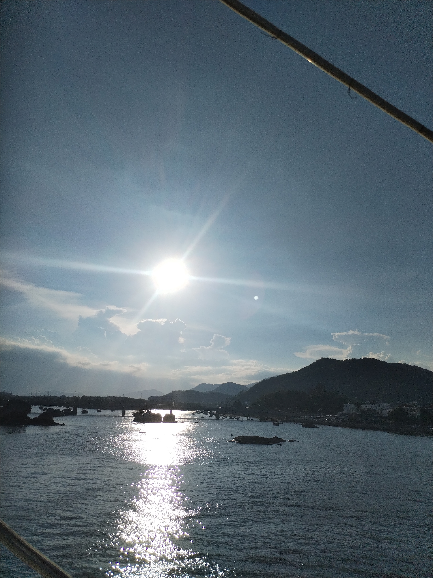 The image depicts a landscape with a large body of water, likely the sea or a wide river, shimmering under the bright sunlight. The sun is prominently positioned in the sky, its rays reflecting strongly on the water surface. In the background, there are silhouettes of hills and a coastline, possibly indicating a coastal or island location. Some structures, potentially buildings, can be discerned along the coastline. In the water, there are a few objects, possibly rocks or small boats. A portion of a metallic structure, such as a railing or pole, is visible in the upper right corner and the bottom left corner of the image, suggesting the photograph was taken from a boat or bridge. Given the location of Nha Trang Ward, Vietnam, it is plausible the water body is part of the South China Sea. The time of day appears to be midday due to the sun's high position and intense glare.